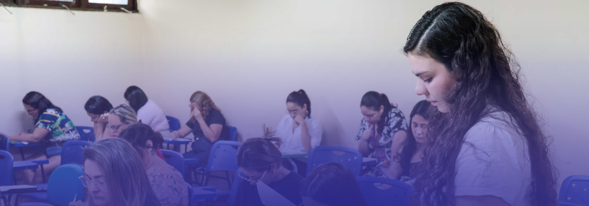 A imagem mostra um grupo de jovens concentrados em um ambiente de sala de aula. Eles estão sentados em carteiras individuais, aparentemente fazendo atividades escritas. As pessoas na cena demonstram foco e dedicação, com expressões de esforço, alguns escrevendo e outros refletindo. O local tem cadeiras azuis e janelas ao fundo. Há uma tonalidade azulada na parte inferior da imagem, possivelmente aplicada como efeito gráfico. O cenário sugere um momento de estudo ou avaliação em grupo, típico de um ambiente