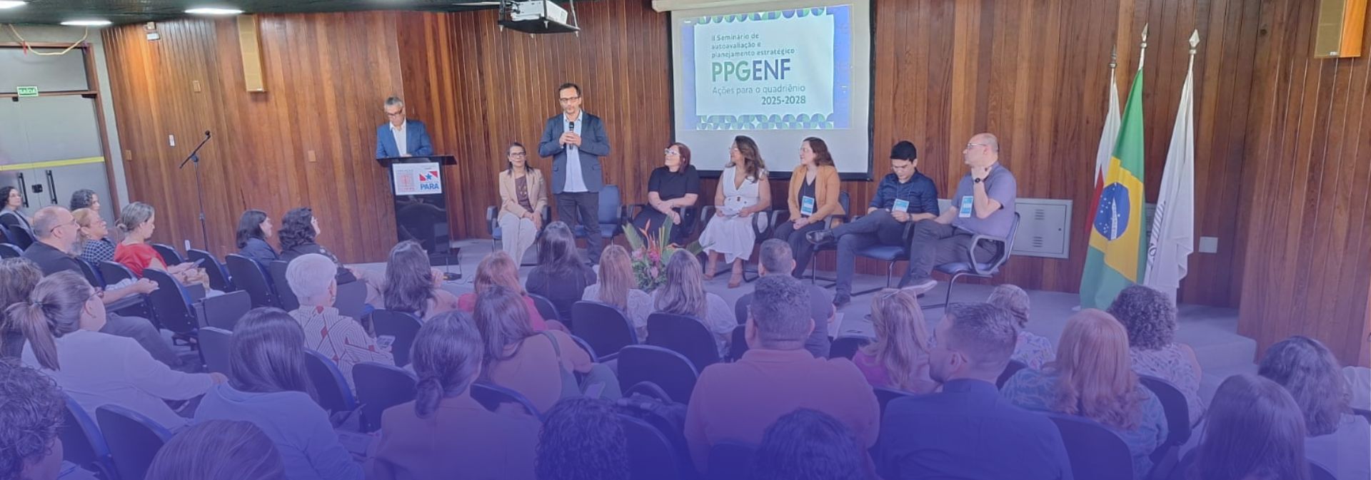 A imagem mostra um ambiente institucional, com pessoas num palco em um auditório com um público assistindo uma palestra. Bandeiras estão ao lado direito do palco.
