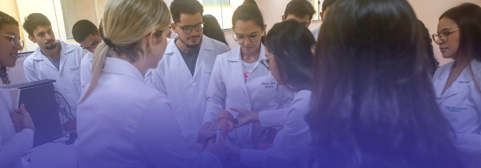 A imagem mostra um grupo de estudantes e docentes do curso de Medicina, vestidos com jalecos, reunidos em uma atividade prática em sala de aula. Sobre a mesa, há um manequim utilizado para treinamento em procedimentos de saúde, enquanto os participantes observam e acompanham a explicação, em um ambiente acadêmico iluminado e organizado para aulas práticas.