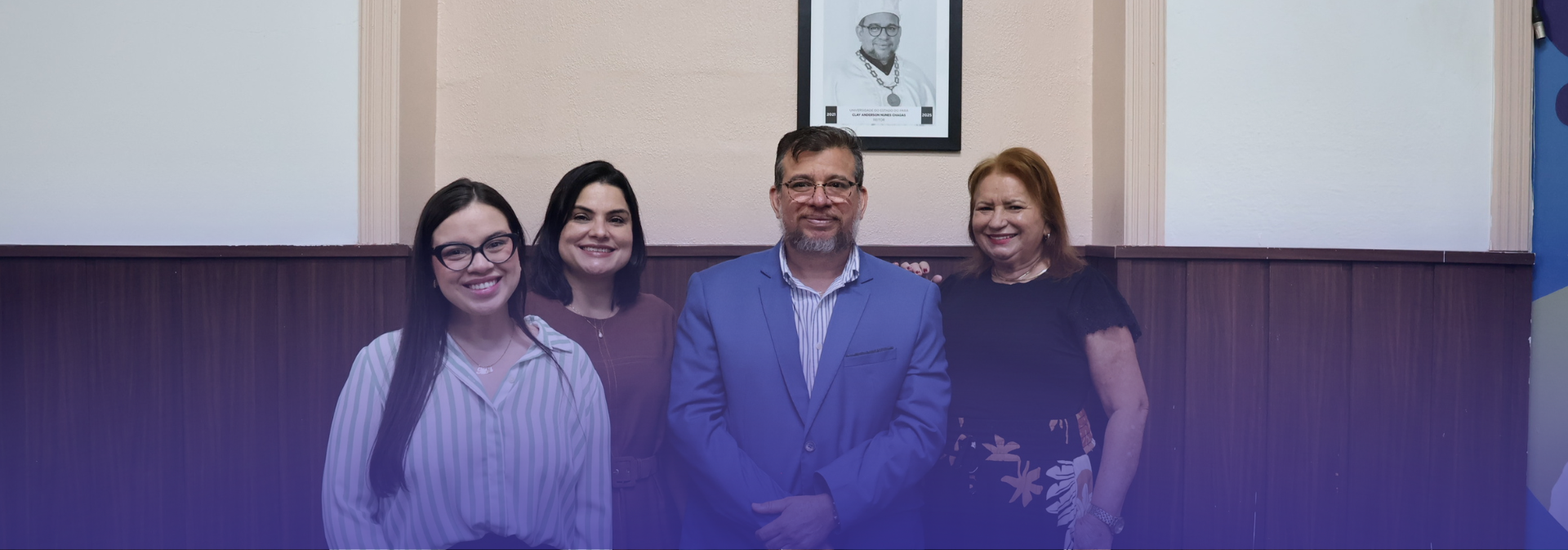 A foto mostra quatro pessoas adultas posando lado a lado em um ambiente institucional interno, todas sorrindo para a câmera. Elas vestem trajes sociais ou casuais formais, transmitindo um clima cordial e profissional. Ao fundo, há uma parede com acabamento em madeira, um retrato emoldurado centralizado acima do grupo e, à direita, parte de um banner institucional.