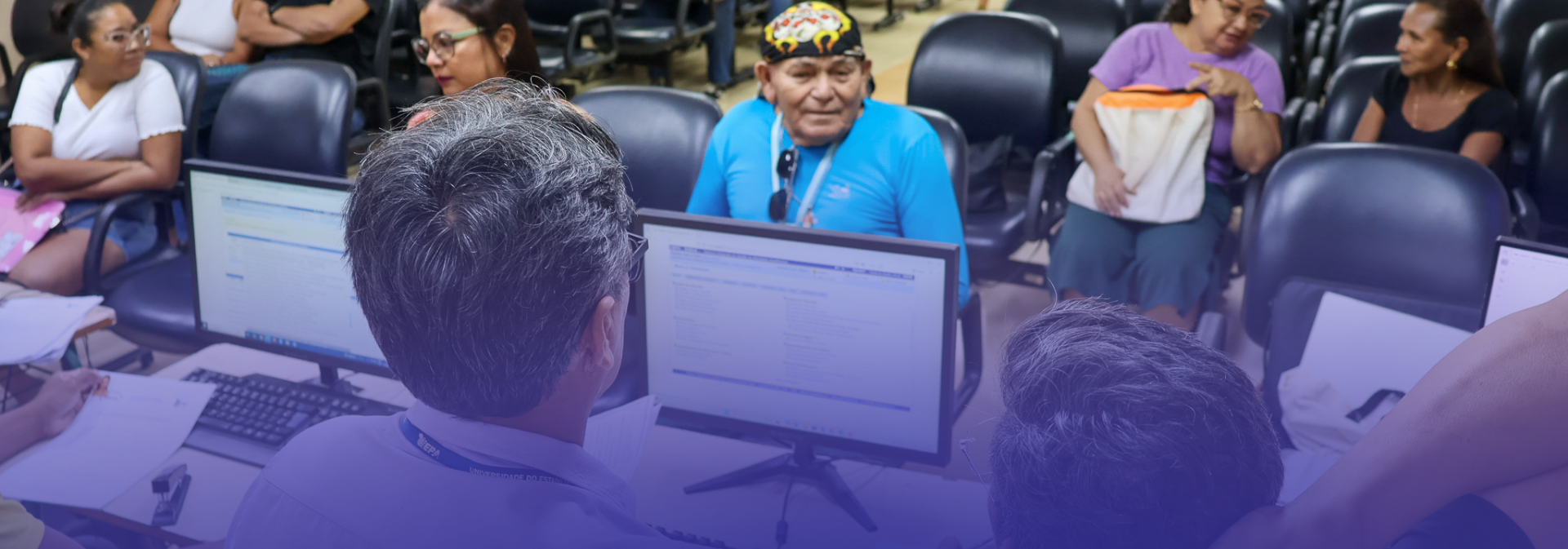 A imagem mostra o atendimento ao público em uma sala com computadores, onde pessoas aguardam e recebem orientação em um ambiente organizado, para a efetivação das matrículas.