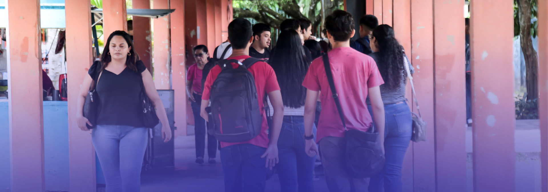 A imagem mostra um corredor de um campus universitário, com várias pessoas — em sua maioria estudantes — caminhando entre colunas alaranjadas, algumas com mochilas e roupas casuais, em um ambiente movimentado e ao ar livre, com árvores ao fundo.