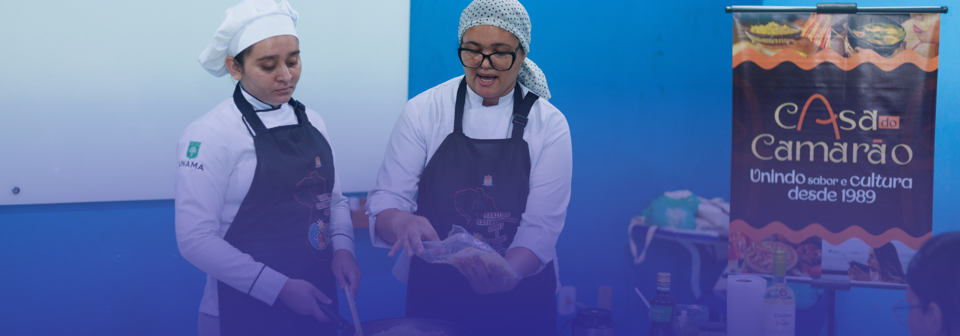 A foto mostra duas mulheres vestidas como chefs, usando aventais e toucas, durante uma atividade culinária. Elas preparam um prato juntas, enquanto uma delas explica o processo, segurando um ingrediente embalado. Ao fundo, há um banner do restaurante “Casa do Camarão”, indicando um ambiente de demonstração ou oficina gastronômica.