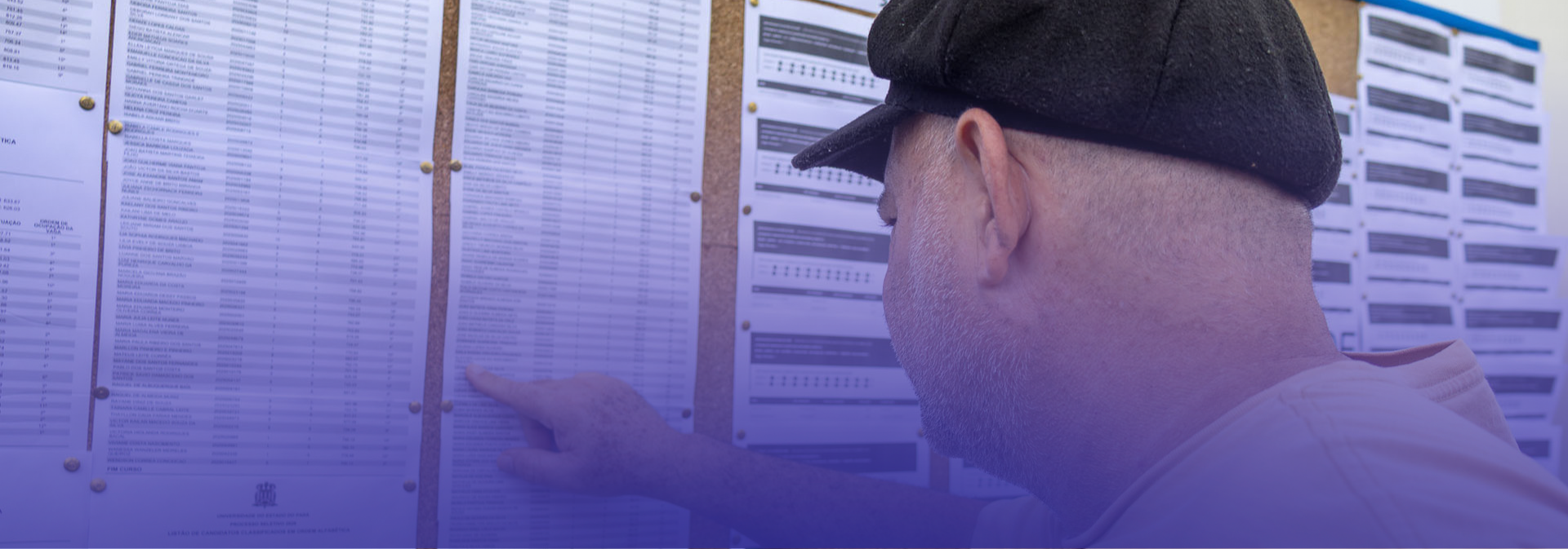 Um homem, visto de perfil e usando um boné preto, observa atentamente uma lista afixada em um mural e aponta para um dos nomes com o dedo, como se estivesse procurando ou confirmando uma informação. Ao fundo, há vários quadros com tabelas.