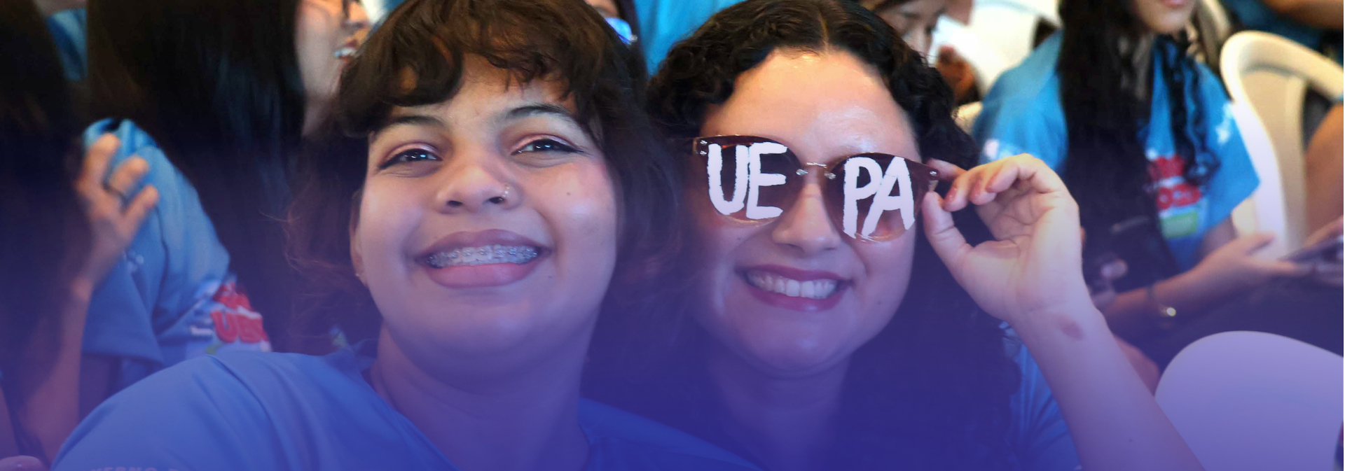 imeiro plano, duas jovens sorriem para a câmera: uma delas usa aparelho ortodôntico e a outra segura um óculos divertido com a inscrição “UEPA” nas lentes, reforçando o sentimento de pertencimento à universidade.