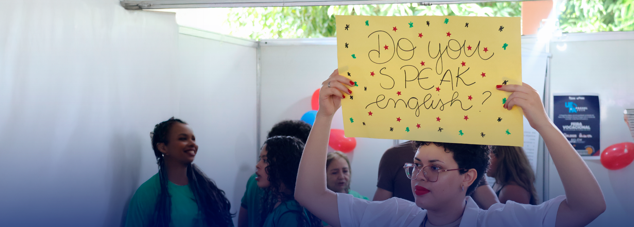 Foto de um ambiente de evento ou feira educacional: em primeiro plano, uma mulher segura um cartaz amarelo com a frase em inglês “Do you speak English?” decorado com pequenas estrelas coloridas. Ao fundo, outras pessoas conversam e há balões e painéis de exposição, sugerindo um estande ou atividade relacionada ao aprendizado de inglês.
