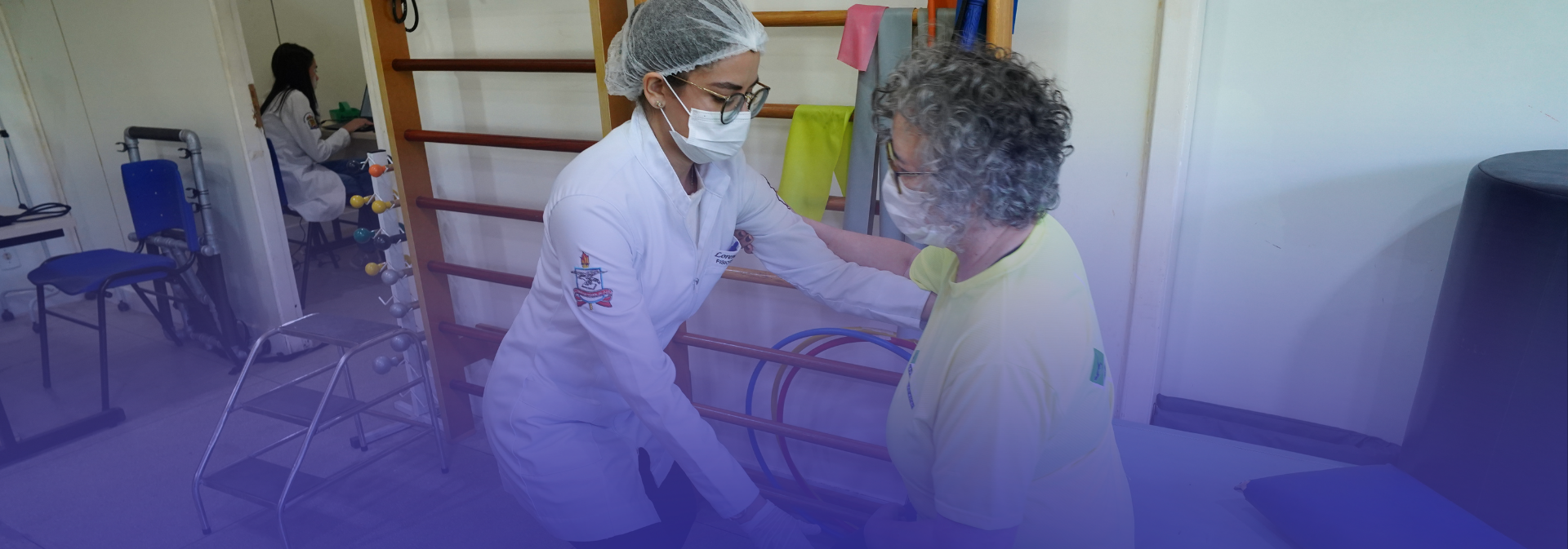 Profissional de saúde, utilizando jaleco, touca e máscara, auxilia uma senhora idosa durante atendimento de fisioterapia ou reabilitação, em ambiente clínico equipado com barras paralelas e materiais terapêuticos. Ao fundo, outra profissional atende um paciente em uma sala adjacente.