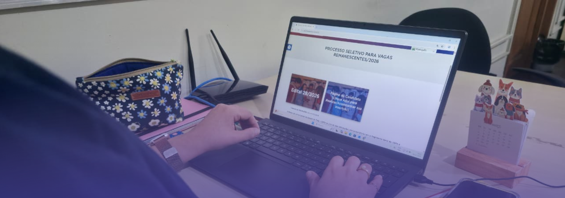 A imagem mostra uma pessoa utilizando um notebook sobre uma mesa de trabalho para acessar uma página de processo seletivo para vagas remanescentes 2026. A pessoa está digitando no computador, enquanto ao redor há objetos de escritório, como um caderno com anotações, um celular, canetas, um estojo floral, um roteador de internet e um pequeno calendário de mesa.