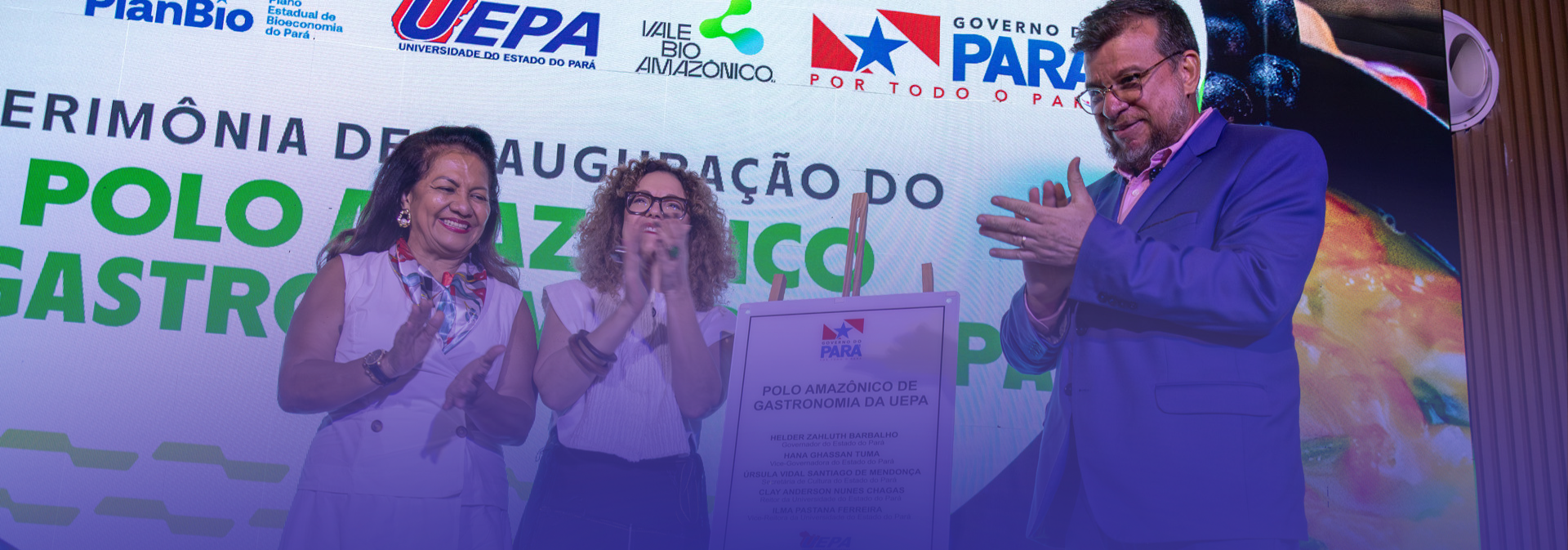 Três pessoas estão em um palco durante uma cerimônia de inauguração. Ao centro, uma placa comemorativa é apresentada enquanto os participantes aplaudem. Ao fundo, um painel exibe os logotipos da Uepa, do Governo do Pará e de parceiros institucionais, além do texto que anuncia a inauguração do Polo Amazônico de Gastronomia. O ambiente é iluminado por luzes de evento e a cena transmite um momento de celebração institucional.