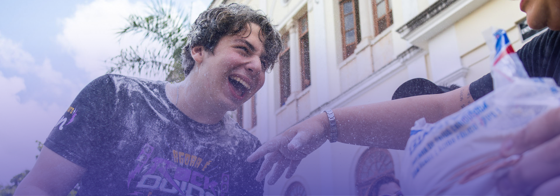 A imagem mostra um jovem sorrindo enquanto é atingido por trigo, em um momento de descontração e celebração ao ar livre. Ao fundo, é possível ver a fachada de um prédio e árvores, enquanto outra pessoa, parcialmente visível, participa da ação, reforçando o clima coletivo e festivo da cena.