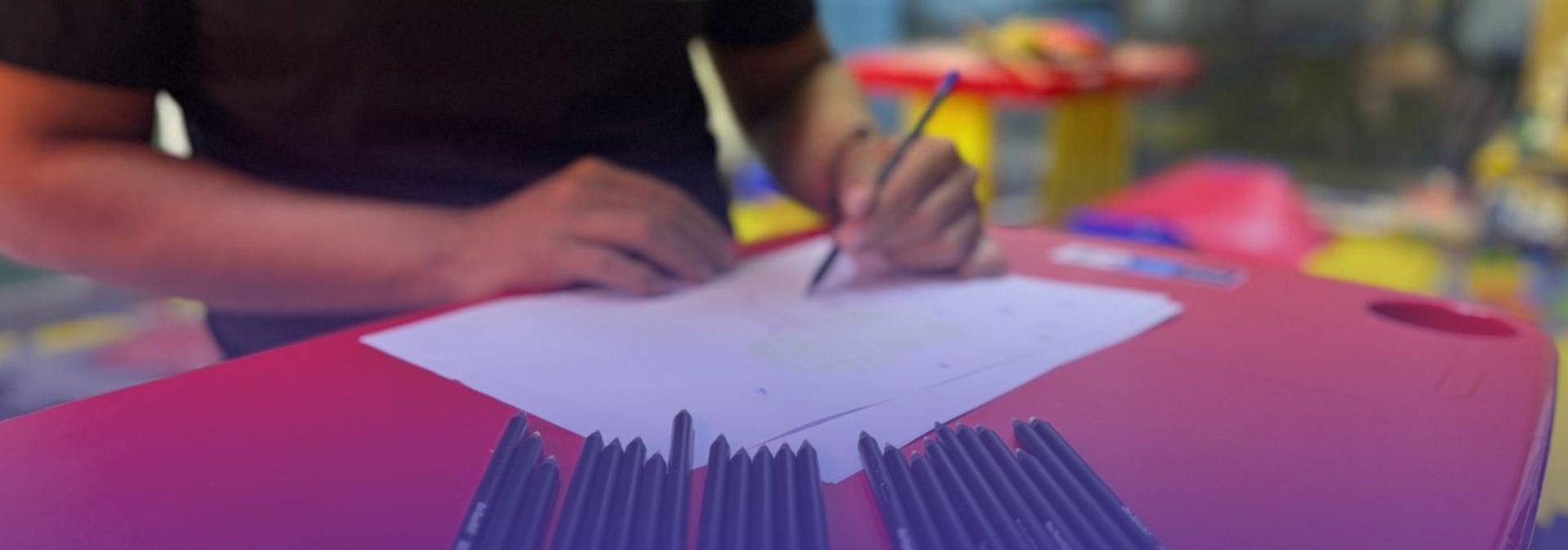 A imagem mostra uma cena de atividade criativa, na Brinquedoteca do CCPPA. Em primeiro plano, sobre uma mesa, há vários lápis de cor organizados lado a lado, apontados em direção ao centro da imagem. Logo acima deles, vê-se uma folha de papel branca onde uma pessoa está desenhando com um lápis.