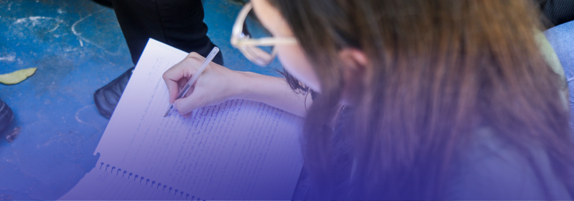 Close-up com filtro azul de pessoa jovem escrevendo em caderno branco. Vê-se parte do rosto com óculos, mãos com caneta e fundo de piso azul. Estilo banner horizontal com desfoque nas bordas.