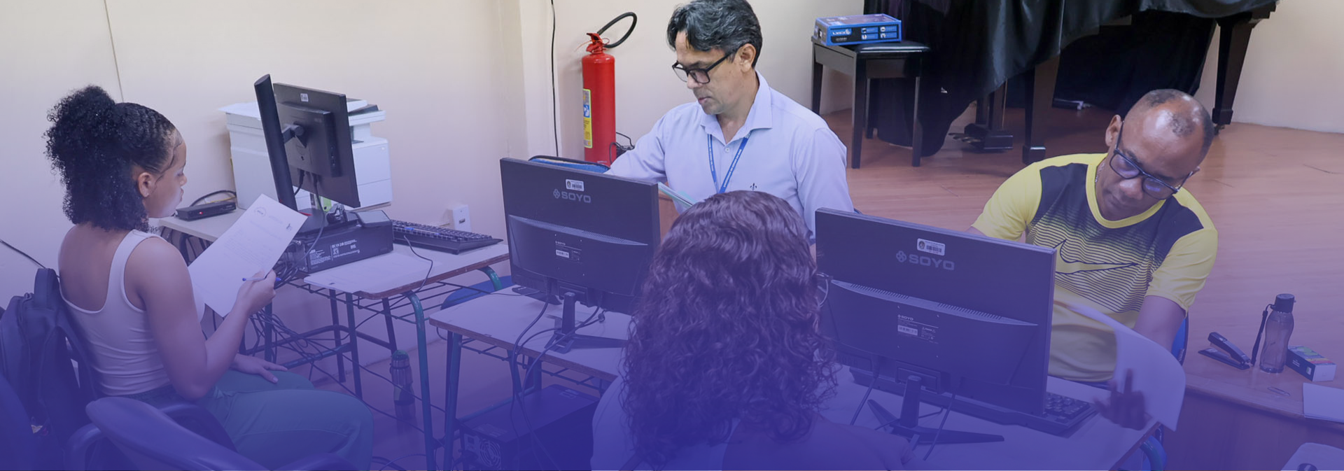 A imagem mostra um ambiente de atendimento ou trabalho administrativo, com quatro pessoas em uma sala equipada com computadores. Duas delas estão sentadas em frente aos monitores, aparentemente realizando atendimentos ou conferindo informações, enquanto outras duas interagem com documentos impressos.
