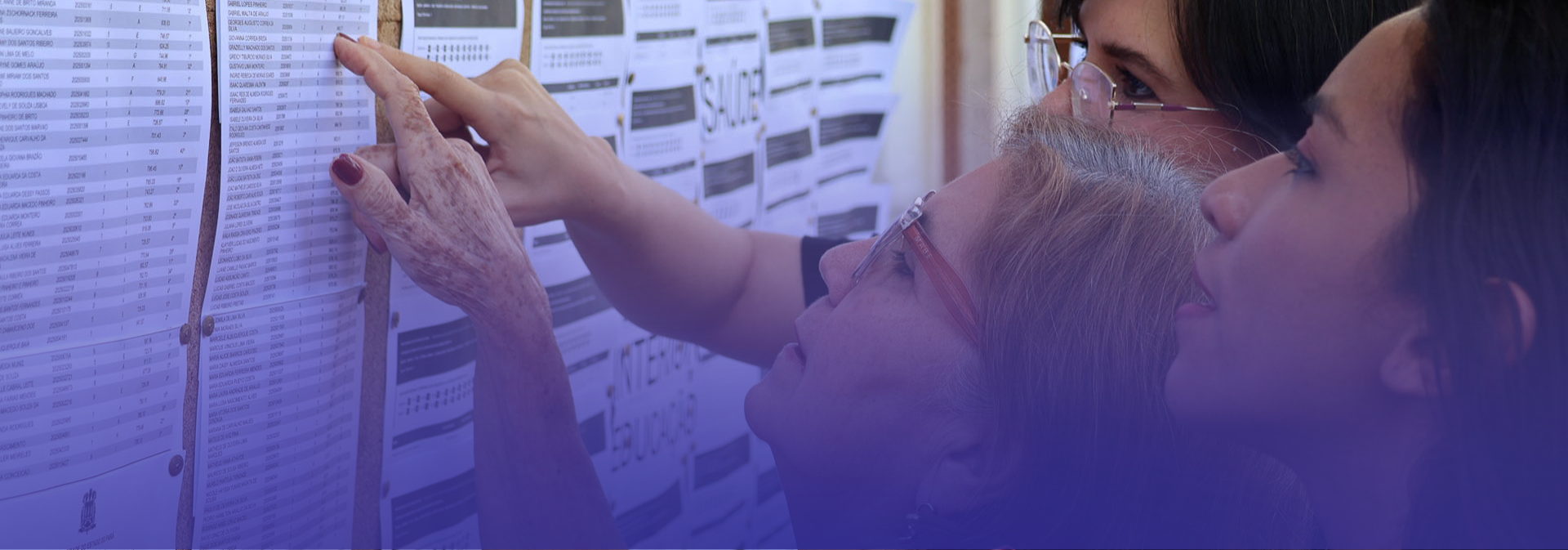 A imagem mostra três mulheres observando atentamente uma lista fixada em um mural. Uma delas aponta com o dedo para um nome ou informação específica no documento, enquanto as outras acompanham a leitura.