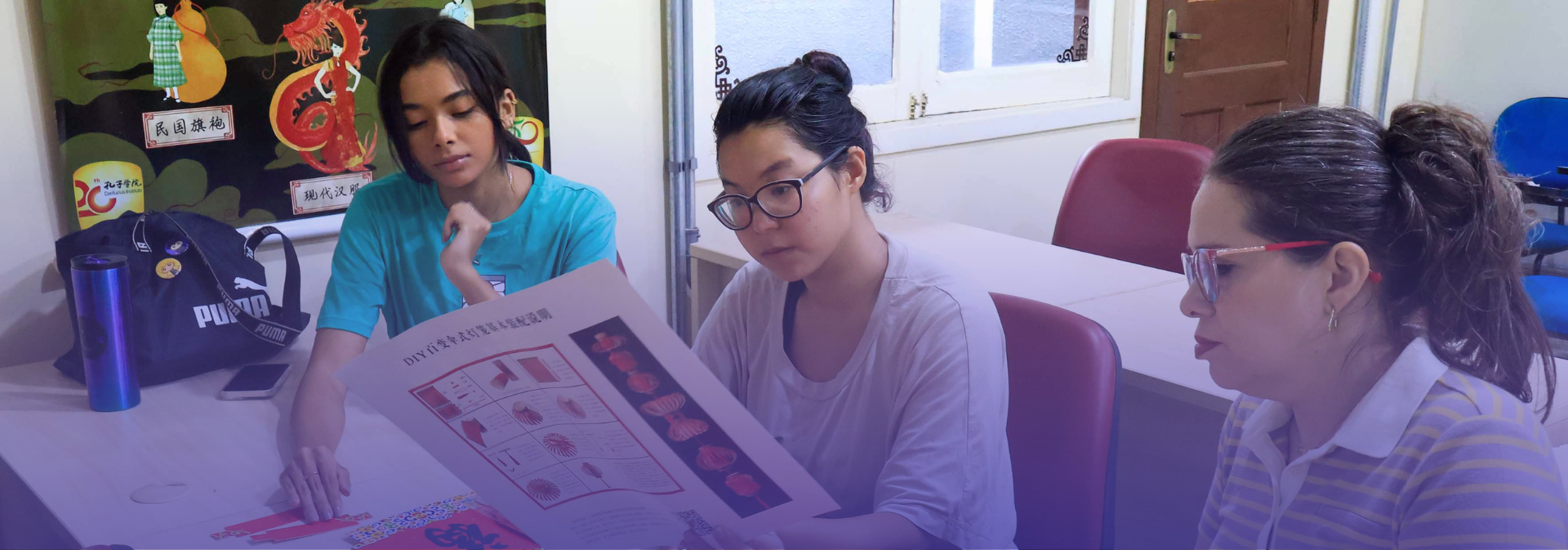 Três pessoas participam de uma oficina cultural, confeccionando artesanato chinês com instruções em mandarim 