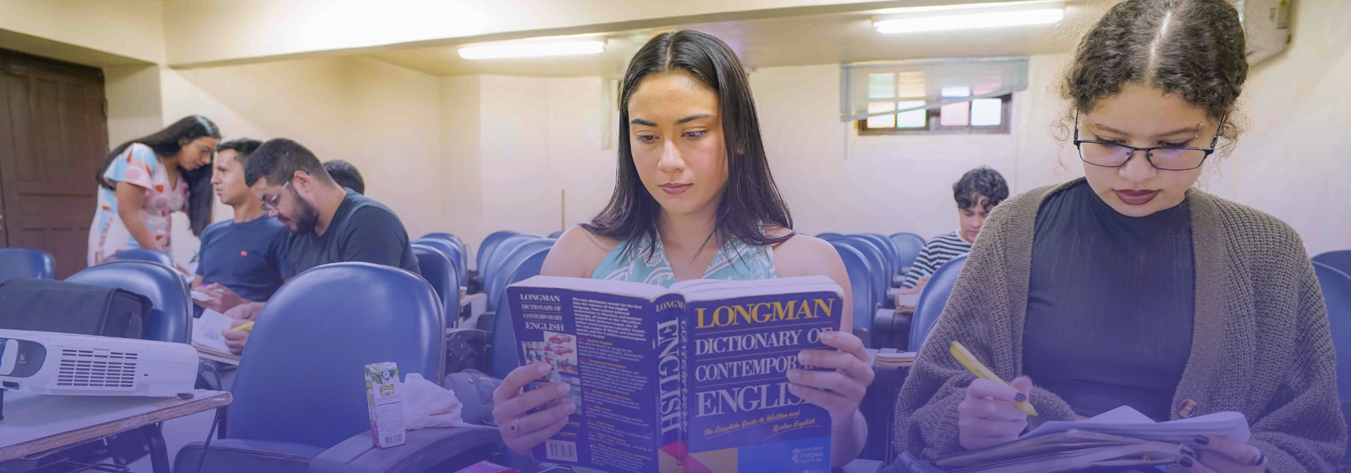 Estudantes sentados em uma sala de aula, com destaque para uma jovem ao centro lendo atentamente um exemplar do dicionário Longman, em Inglês