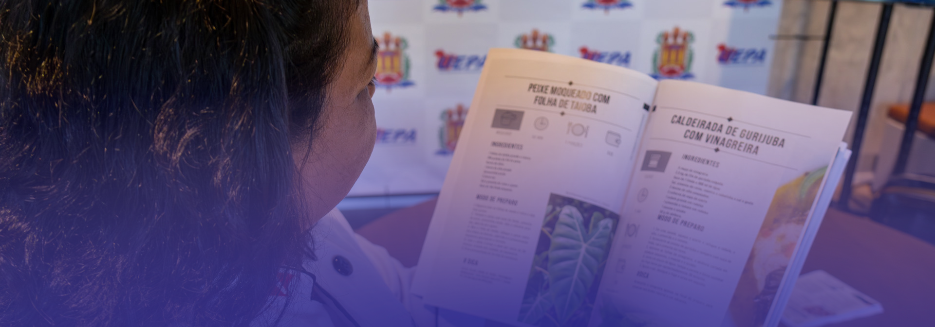 Uma mulher folheia um livro de receitas enquanto observa atentamente as páginas abertas, que apresentam preparações com ingredientes regionais, como folhas e pratos típicos. A cena ocorre em um ambiente institucional, com um fundo contendo a identidade visual da Uepa, e sobre a mesa há outros materiais impressos relacionados à gastronomia e às PANCs.