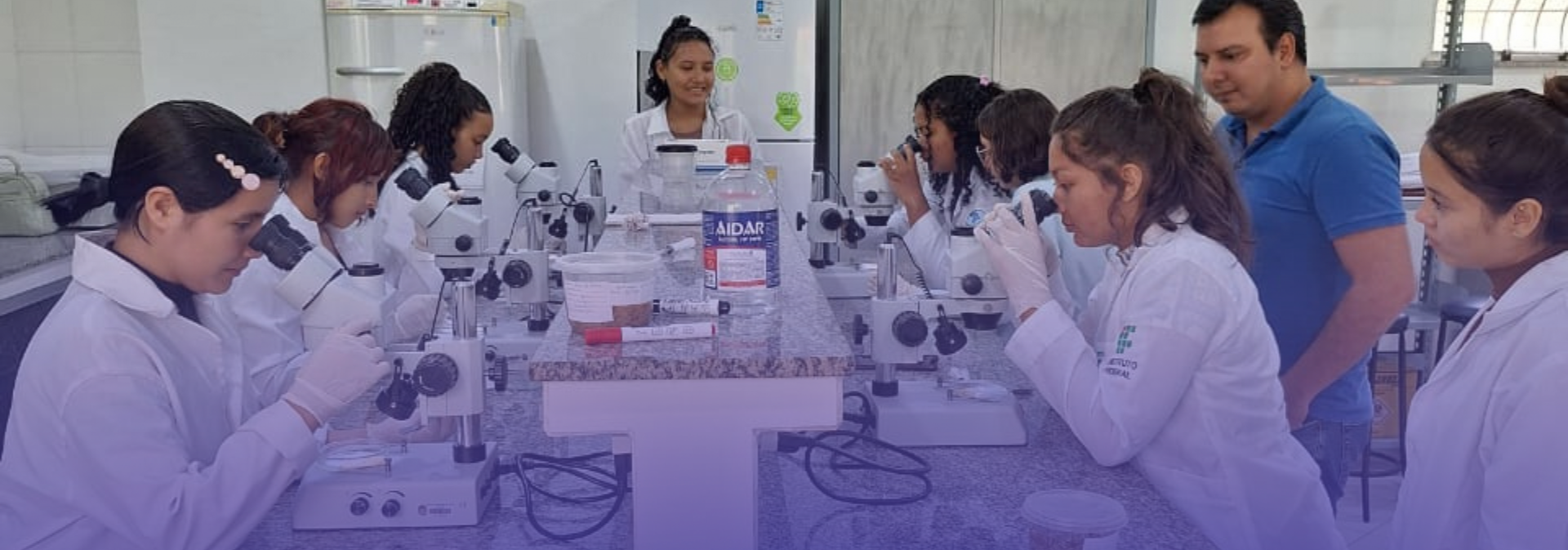Vários estudantes e um instrutor estão em um laboratório, com a maioria dos alunos usando jalecos e olhando através de microscópios.