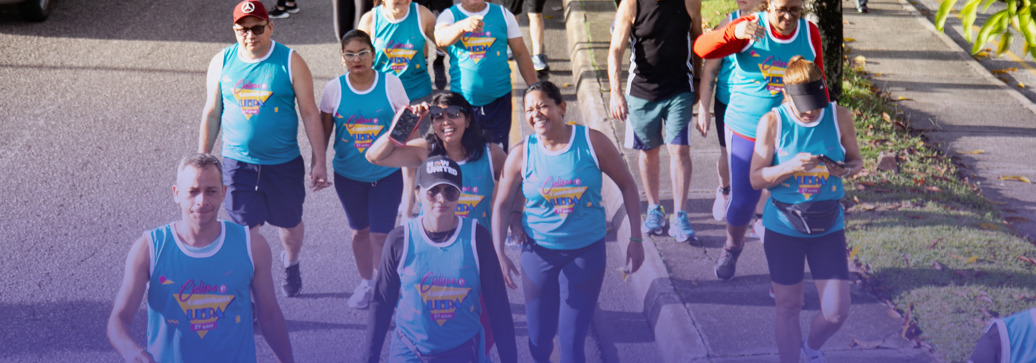 A imagem mostra um grupo de pessoas participando de uma atividade ao ar livre em uma via urbana, possivelmente uma corrida ou caminhada. Os participantes usam camisetas esportivas semelhantes, em tom azul, sugerindo que fazem parte de um mesmo evento. 