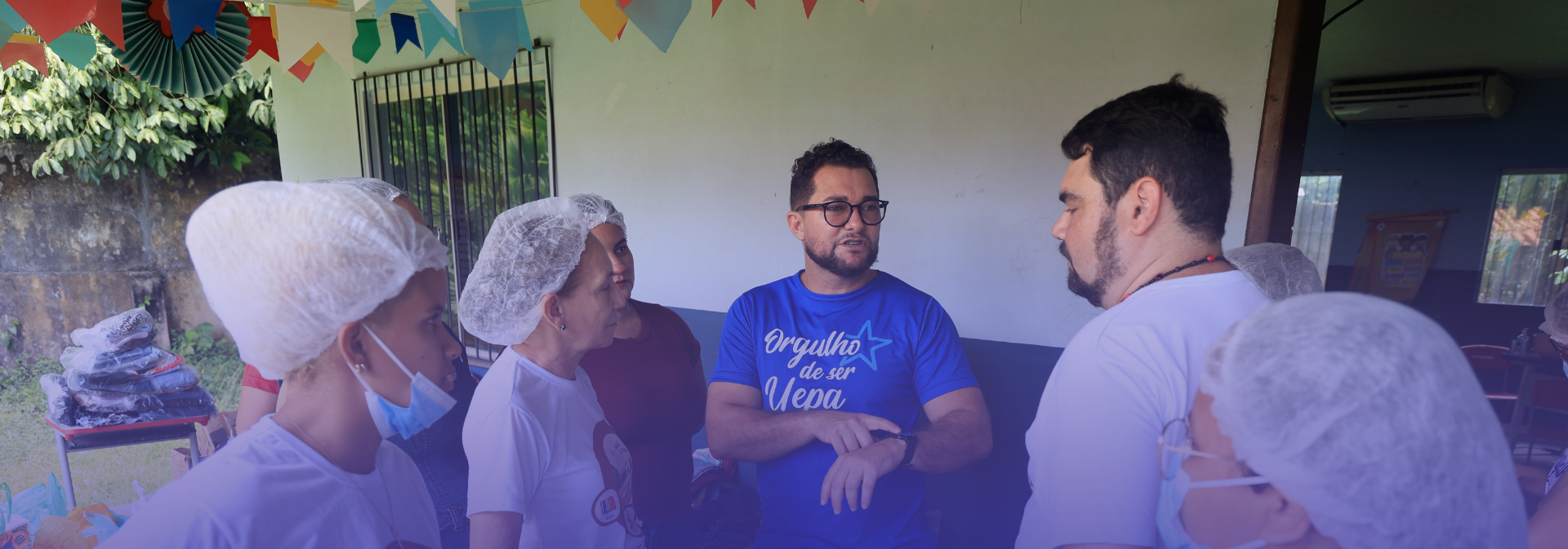 Grupo de pessoas reunidas em um ambiente coberto, possivelmente em uma ação comunitária ou atividade coletiva. A maioria usa toucas descartáveis e algumas também utilizam máscaras, sugerindo manipulação de alimentos. No centro, um homem com camiseta azul conversa e gesticula com os demais, que o escutam atentamente. O espaço está decorado com bandeirinhas coloridas ao fundo.