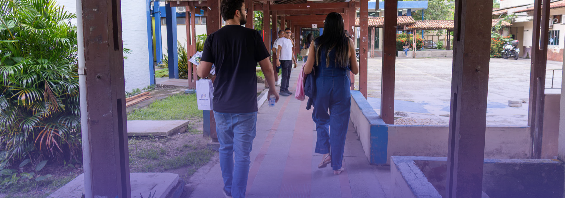 Duas pessoas caminham por um corredor coberto em um ambiente que parece ser um campus ou instituição, com estruturas de madeira, áreas verdes ao lado e outras pessoas ao fundo. Uma delas carrega uma sacola e uma garrafa de água, enquanto a outra segura alguns objetos e veste um macacão azul.