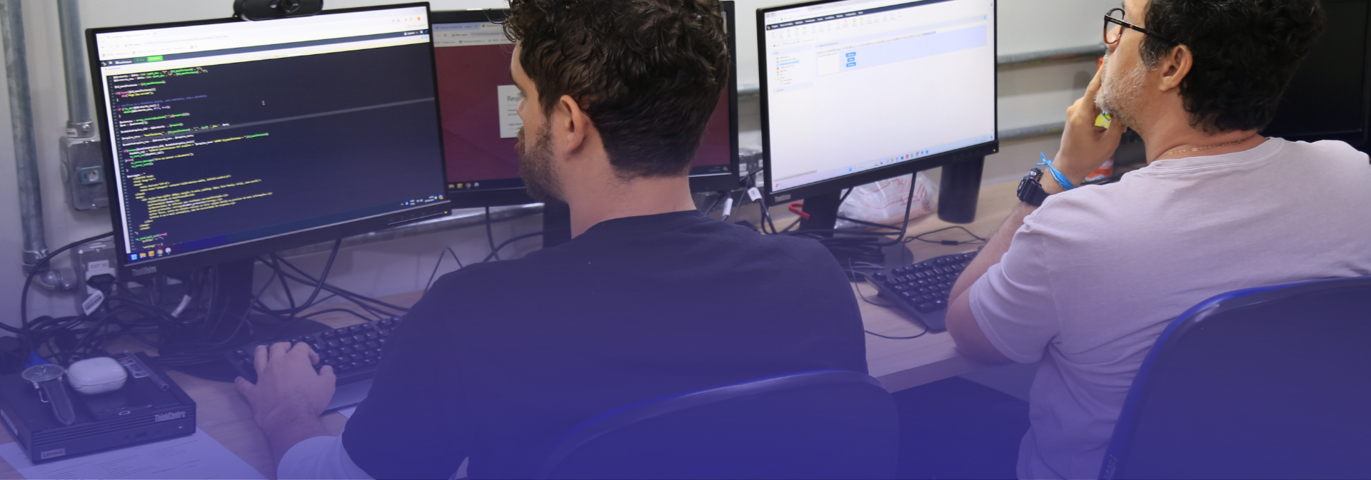 A imagem mostra dois homens sentados lado a lado em um ambiente de escritório, trabalhando em computadores. Um deles digita enquanto observa códigos exibidos na tela, e o outro parece concentrado, apoiando a mão no rosto diante de seu monitor. Há múltiplos monitores, teclados e cabos sobre a mesa, indicando um contexto de trabalho ligado à programação ou tecnologia.