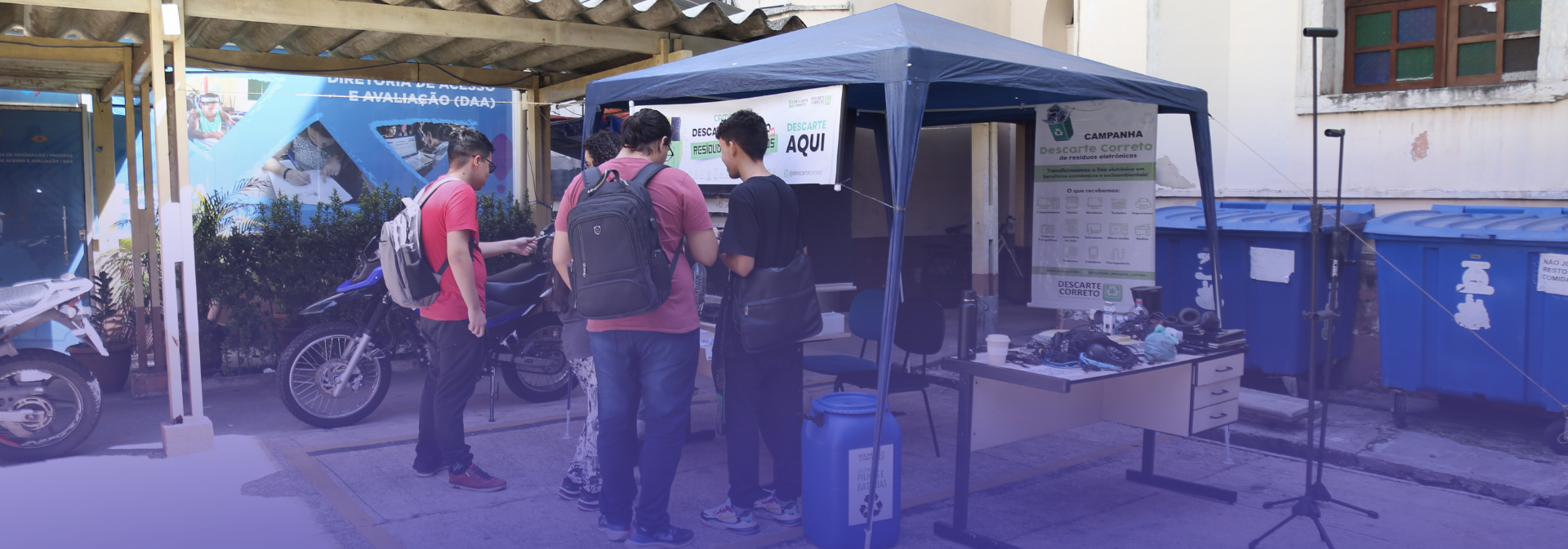 A imagem mostra uma ação de coleta de resíduos eletrônicos realizada em uma área externa de uma instituição. Sob uma tenda azul, algumas pessoas observam e realizam o descarte de equipamentos eletrônicos, como cabos e periféricos.