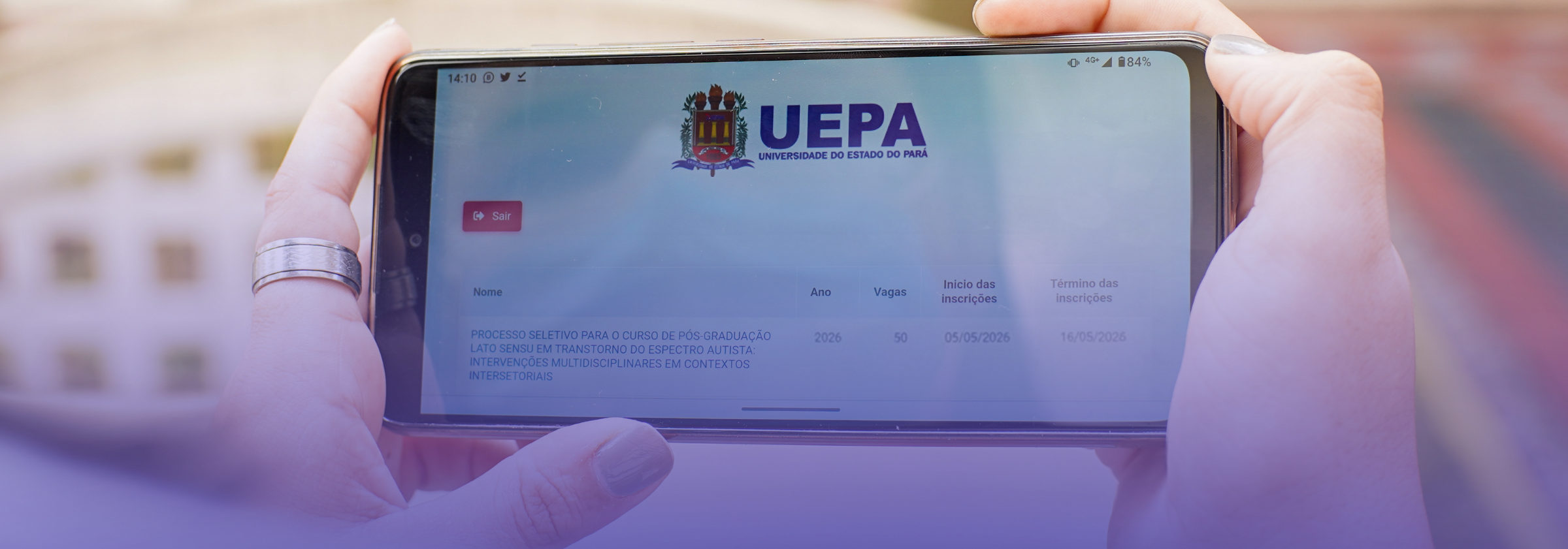 Pessoa segura um smartphone na horizontal, com as duas mãos, acessando uma página da Universidade do Estado do Pará. Na tela, aparece um sistema de inscrição com informações sobre um processo seletivo de pós-graduação, incluindo ano, número de vagas e período de inscrição. O fundo está desfocado e há uma sobreposição em tom azulado na imagem.