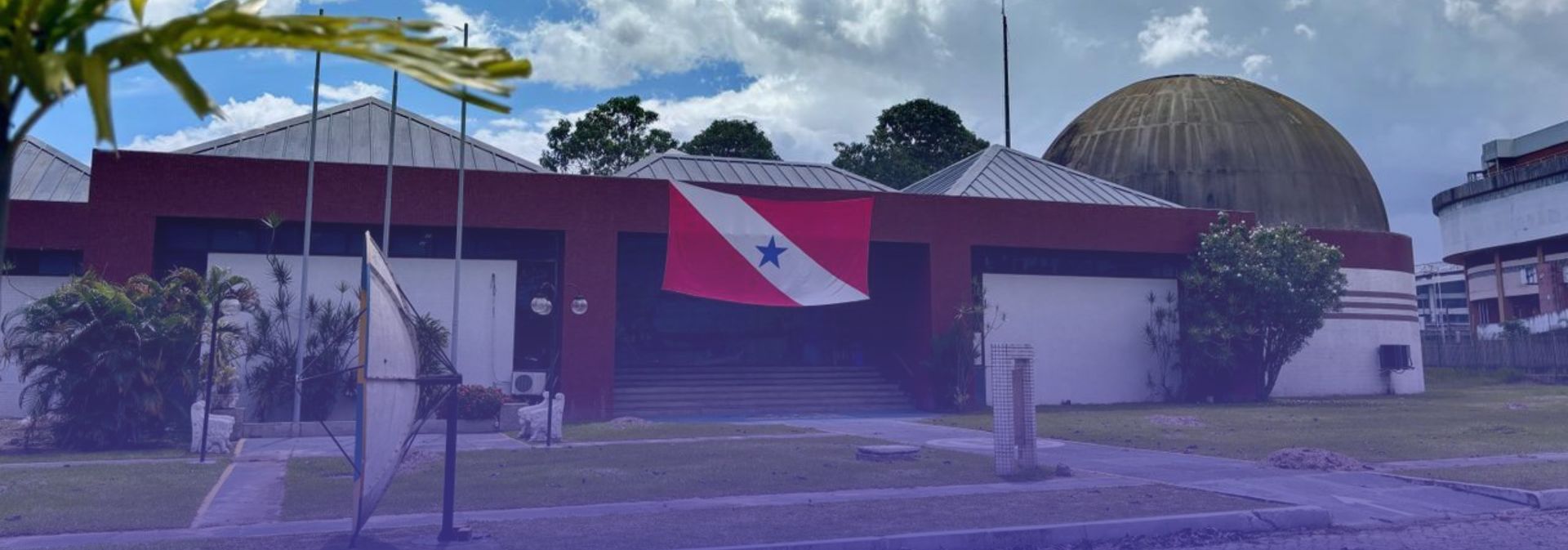 A imagem mostra a área externa do Centro de Ciências e Planetário do Pará, em um dia de céu azul com grandes nuvens brancas ao fundo. Em destaque, aparece a fachada do prédio, marcada por tons de vermelho escuro e branco, além da tradicional cúpula do planetário ao lado direito da construção.