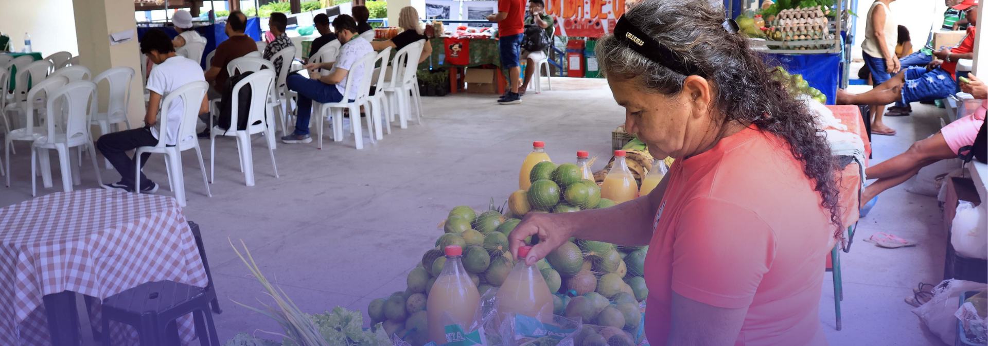 Fotografia mostra uma feira realizada em espaço coberto, com bancas de produtos regionais ao fundo e pessoas sentadas em cadeiras plásticas acompanhando uma atividade. Em primeiro plano, uma mulher de cabelos grisalhos, presa com tiara preta, organiza garrafas de suco e frutas cítricas expostas em uma banca. Ao redor, há verduras, legumes e outros produtos comercializados no local.