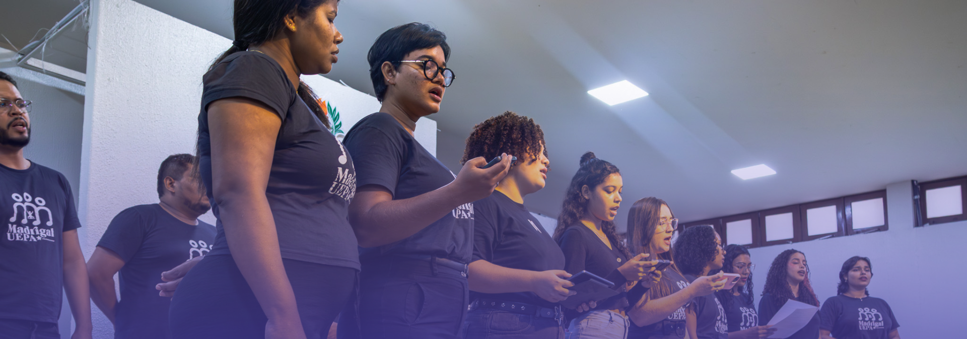 Integrantes do Madrigal da Uepa se apresentam em grupo durante atividade cultural, cantando em ambiente interno e segurando celulares e folhas de apoio para acompanhar a apresentação musical.