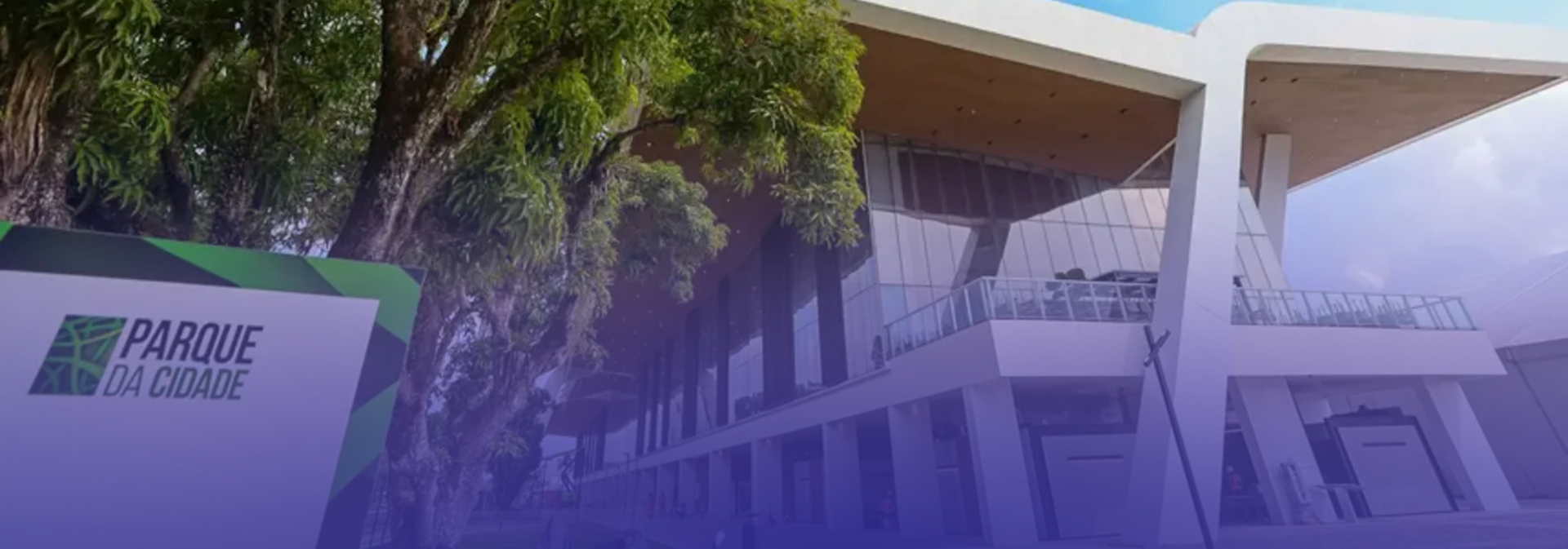Imagem externa do Parque da Cidade, com destaque para a arquitetura moderna do prédio, marcada por grandes estruturas brancas, fachadas envidraçadas e amplos espaços abertos. À esquerda, há uma placa de identificação do local, enquanto árvores de grande porte complementam a paisagem urbana e arborizada.
