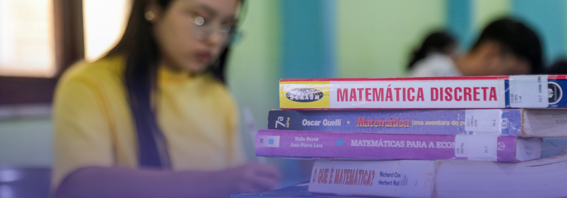 Estudante desfocada em primeiro plano escreve em uma sala de aula enquanto, em destaque à direita da imagem, uma pilha de livros de matemática repousa sobre uma mesa. Entre os títulos visíveis estão obras sobre matemática discreta, ensino da matemática e aplicações da matemática à economia. Ao fundo, outros estudantes aparecem desfocados em ambiente de estudo, reforçando o contexto acadêmico e de formação na área.