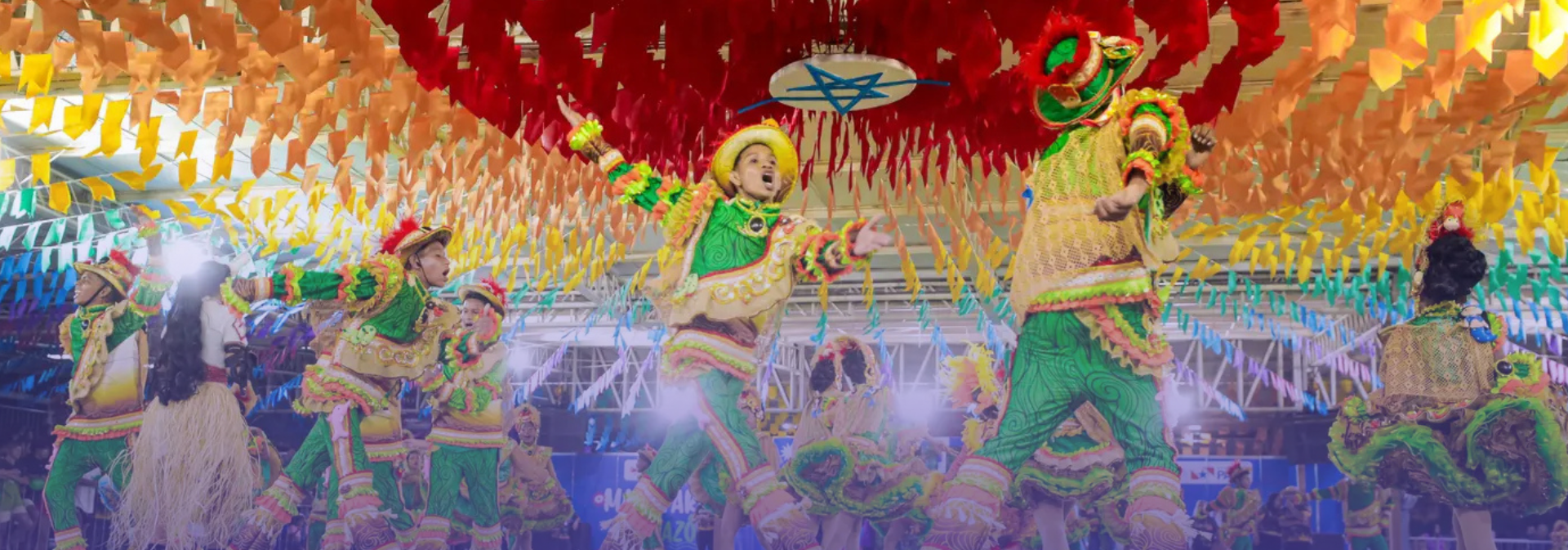 Integrantes de uma quadrilha junina se apresentam em um arraial decorado com centenas de bandeirolas coloridas suspensas no teto. Em primeiro plano, dançarinos vestem trajes típicos em tons de verde, amarelo e laranja, com chapéus e adereços coloridos, enquanto executam uma coreografia. Ao fundo, outros participantes completam a apresentação sob iluminação cênica, destacando o clima festivo e a valorização das tradições culturais do período junino.