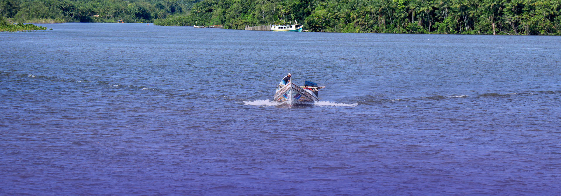 Uma pequena embarcação navega por um amplo rio amazônico, cercado por densa vegetação às margens. Ao fundo, outras embarcações estão ancoradas, compondo uma paisagem típica dos rios da Amazônia.