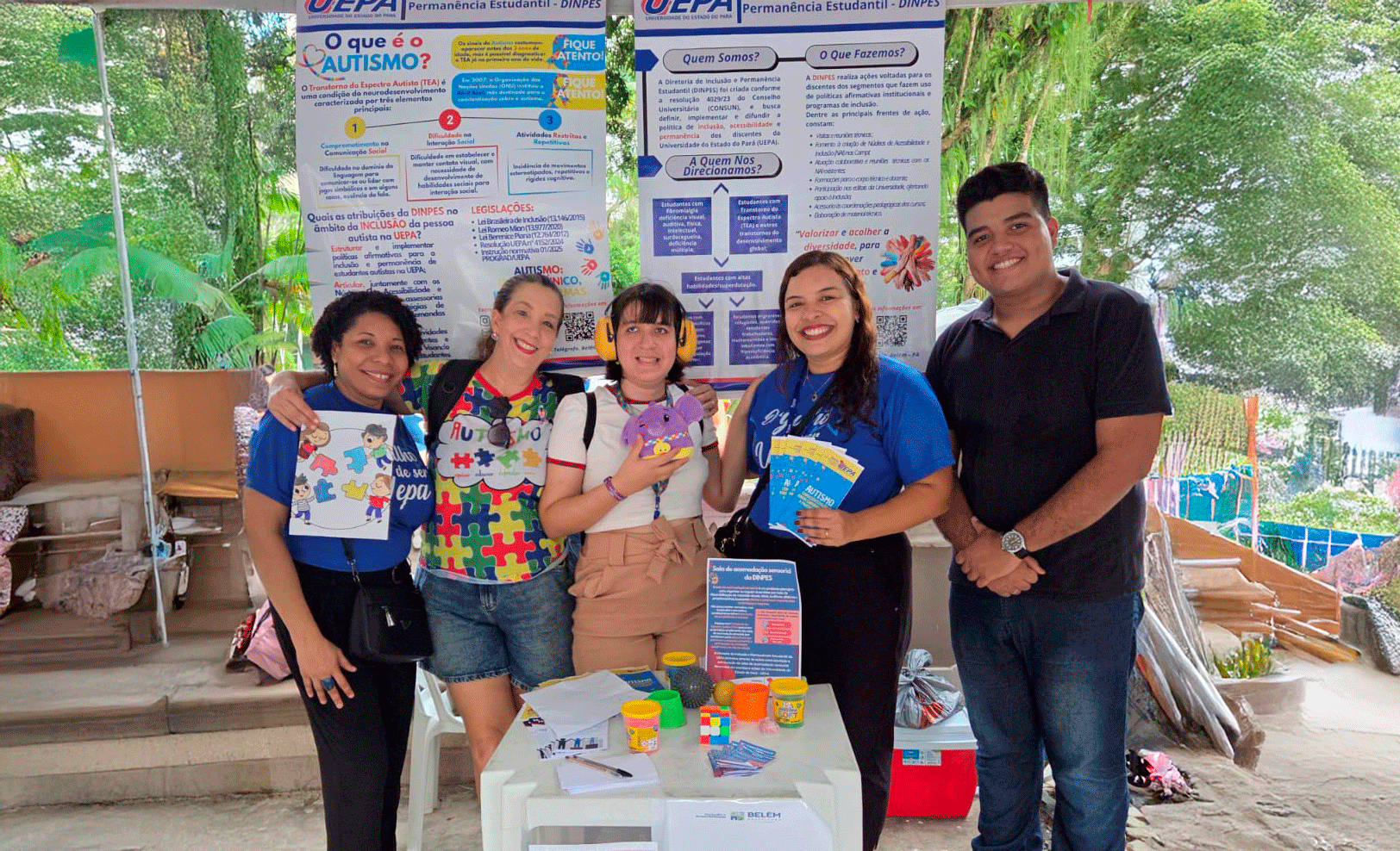 Cinco pessoas estão à frente de de cartazes informativos sobre inclusão e autismo, segurando folhetos e materiais temáticos em um ambiente externo.