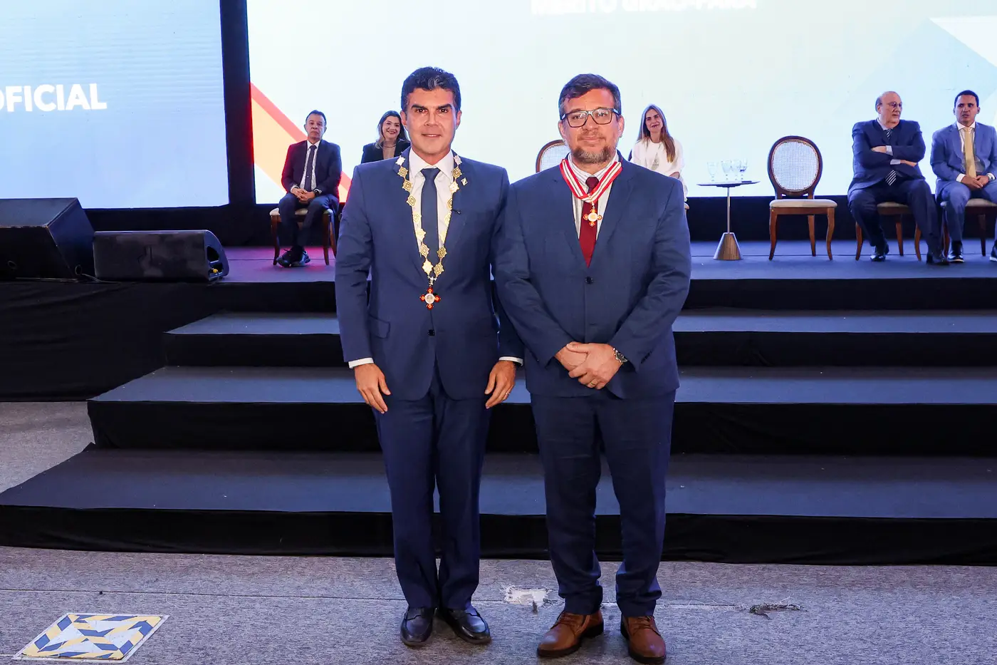 Dois homens em trajes formais posam lado a lado em um palco durante uma cerimônia oficial. Ambos vestem ternos escuros, sendo que um deles usa uma insígnia com colar cerimonial e o outro ostenta uma medalha no peito. Ao fundo, há outras autoridades sentadas em cadeiras e um telão iluminado, compondo o cenário de um evento institucional solene.