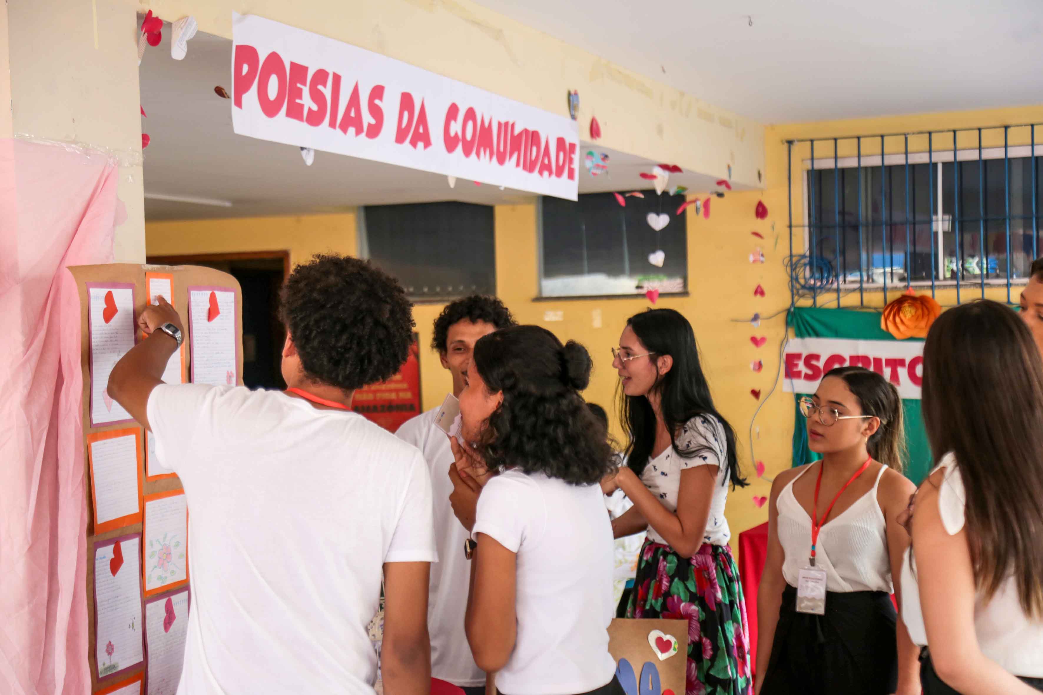 Na imagem, um grupo de jovens participa de uma atividade intitulada “Poesias da Comunidade”, durante um evento escolar ou universitário. Eles observam e comentam textos expostos em painéis de papel pardo, decorados com bordas coloridas e pequenos corações recortados. O ambiente está enfeitado com fitas e corações pendurados no teto, criando um clima acolhedor e afetivo. Ao fundo, há uma faixa com a palavra “ESCRITO” parcialmente visível, sugerindo um espaço dedicado à escrita e à leitura coletiva.