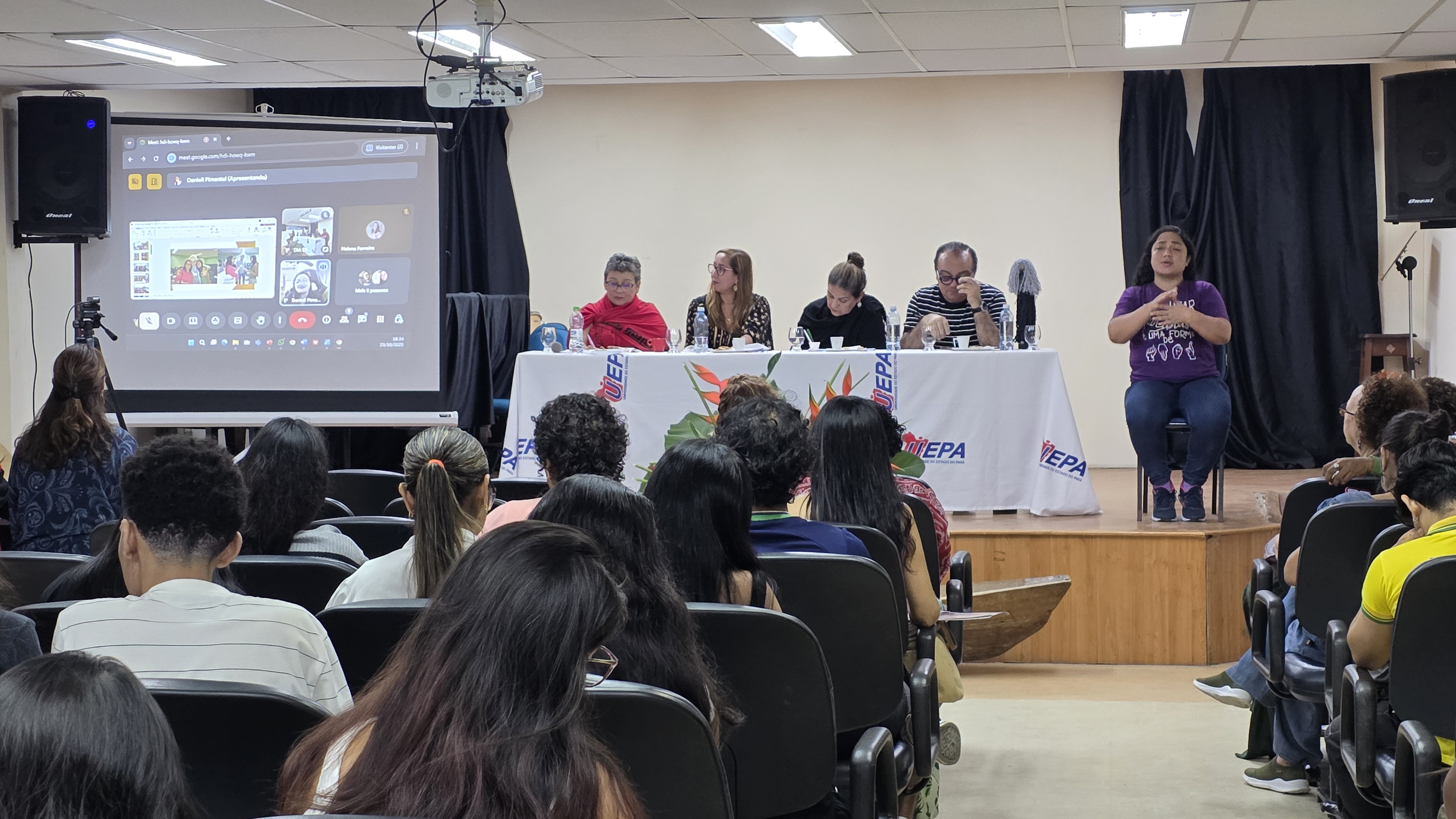 XX Seminário do Cuma na Sala de Recitais do CCSE/UEPA com público atento a uma mesa composta por cinco pessoas, entre elas uma intérprete de Libras. À esquerda, um telão exibe participantes em videoconferência, integrando o evento de forma híbrida. Sobre a mesa, há toalhas com a logomarca da Uepa e arranjo floral ao centro. A cena retrata um momento de debate acadêmico com acessibilidade e participação presencial e online.
