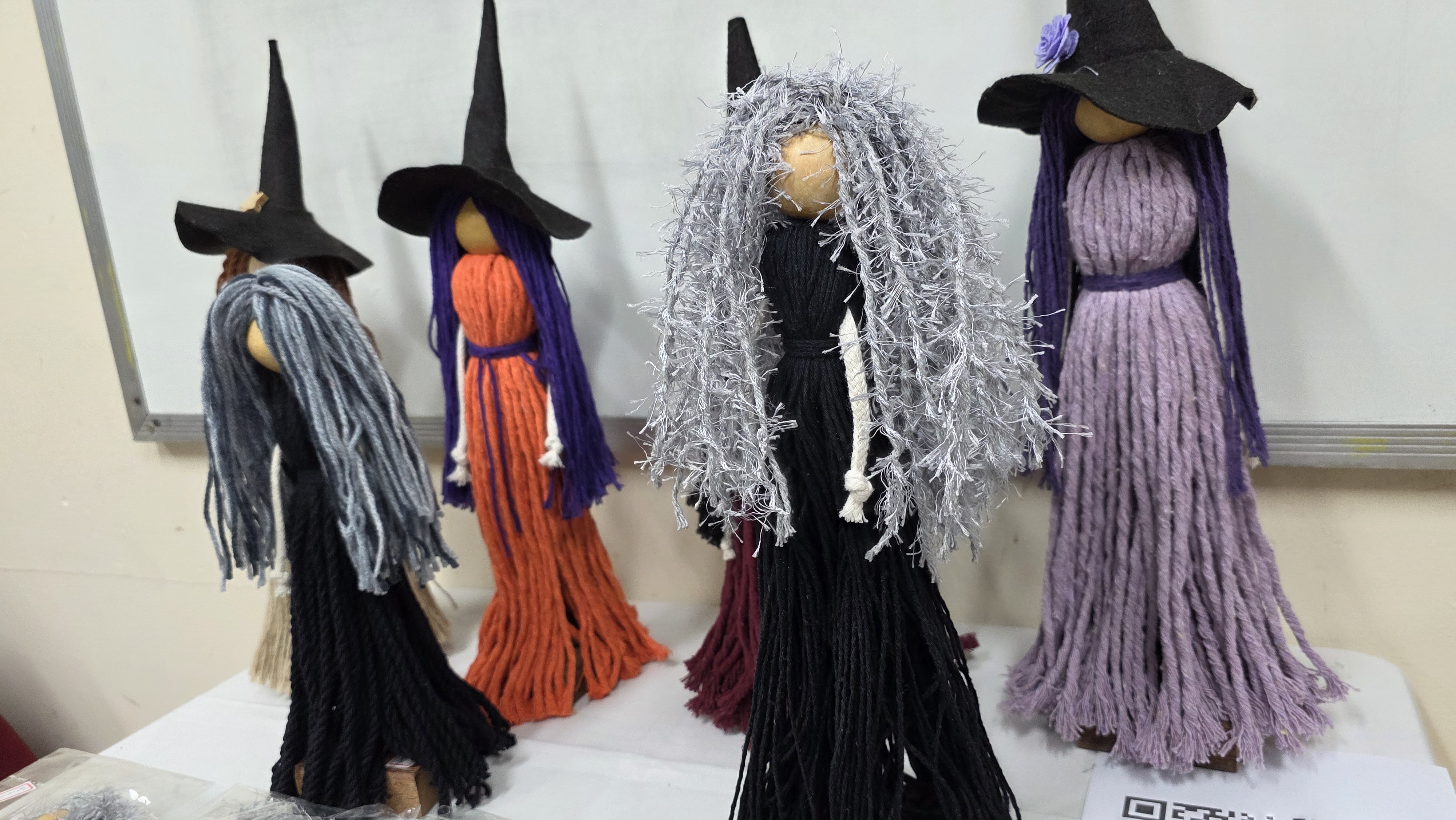 Bonecas artesanais feitas com fios de lã coloridos e chapéus de bruxa pretos estão expostas sobre uma mesa branca, em frente a um quadro branco. Cada boneca tem um visual único, com variações nas cores das roupas e dos cabelos — em tons de cinza, preto, roxo e laranja — lembrando figuras místicas ou bruxas. A imagem destaca o cuidado artesanal e o aspecto criativo das peças.