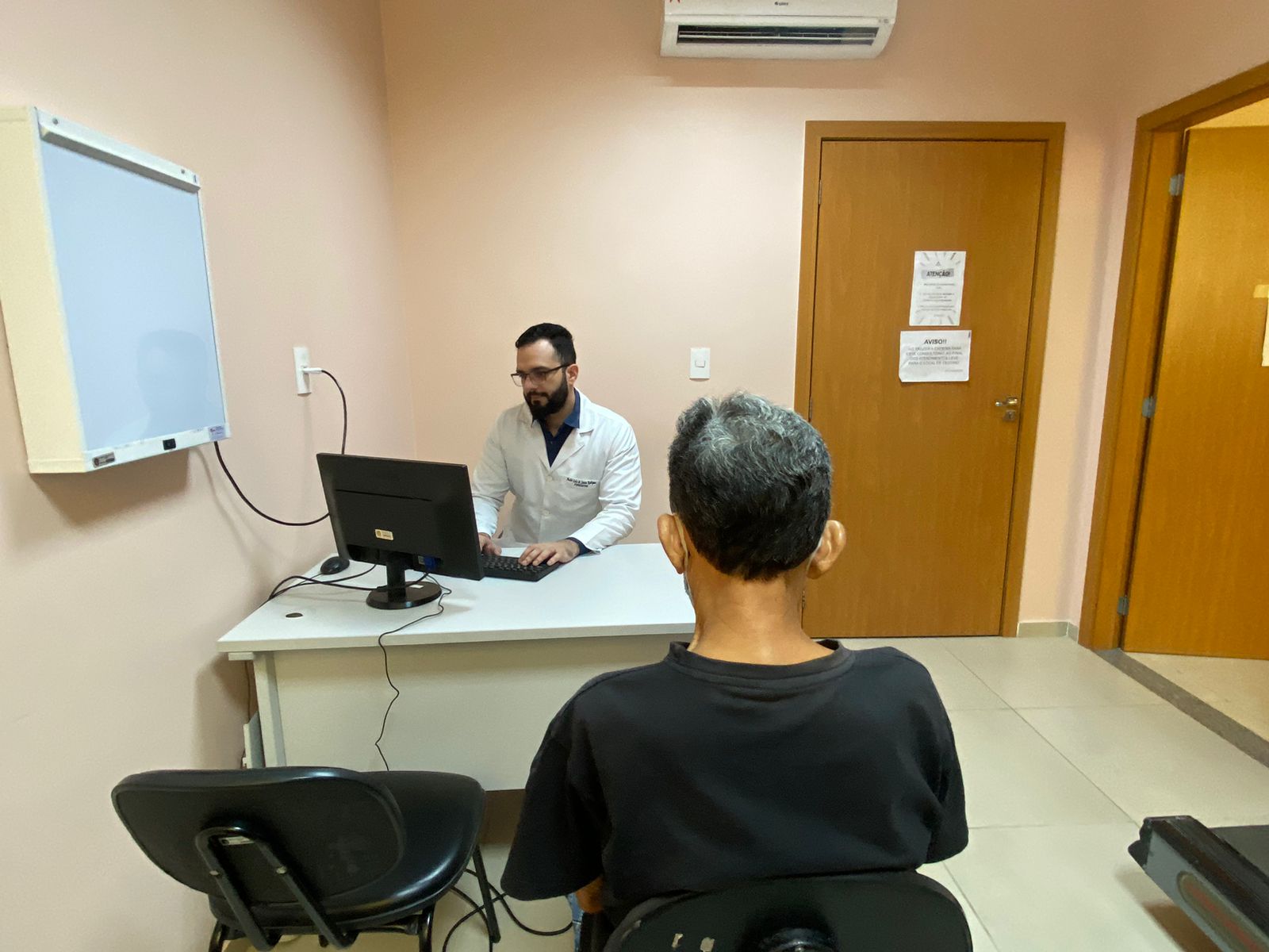 A imagem mostra um consultório médico, onde um profissional de saúde de jaleco branco está sentado à mesa usando um computador, enquanto um paciente, de máscara, está sentado de frente para ele.