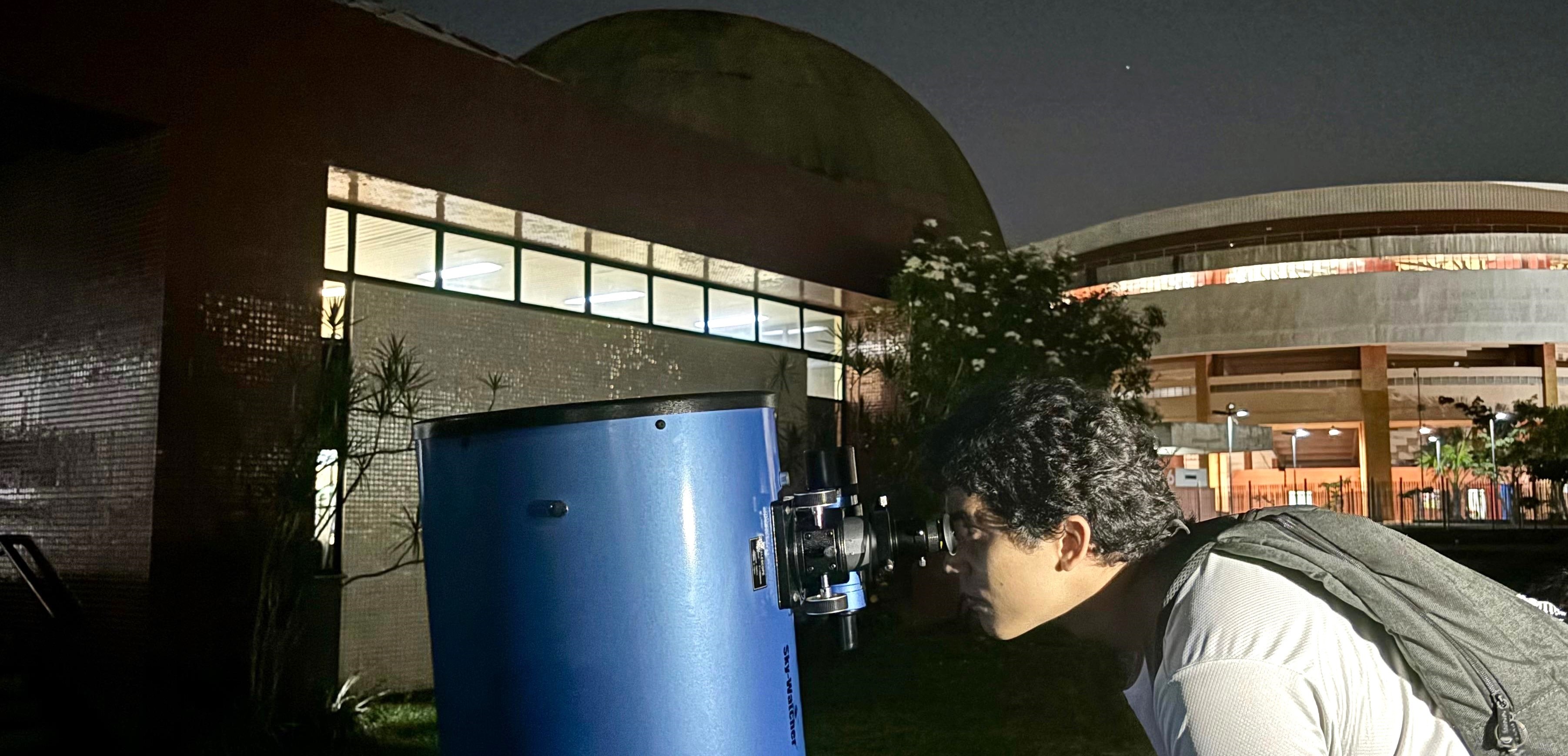 A imagem mostra um jovem observando o céu noturno por meio de um telescópio azul. Ele está inclinado para frente, olhando pela ocular do equipamento, e usa uma mochila cinza nas costas e uma camiseta clara. A cena ocorre em frente ao prédio do Centro de Ciências e Planetário do Pará.