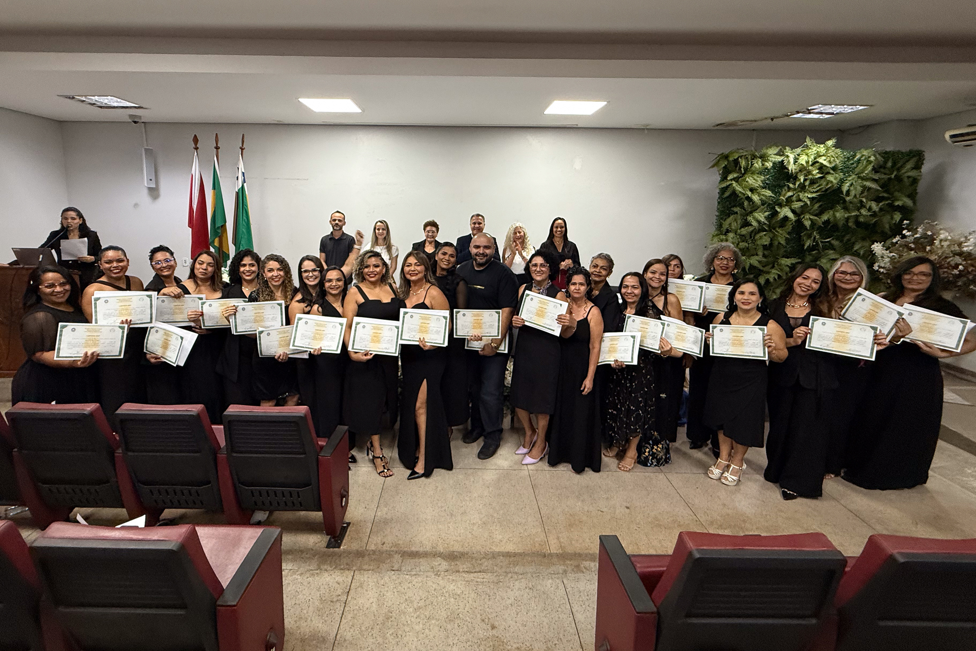 A imagem mostra um grupo numeroso de pessoas posando para uma foto em um auditório. A maioria está vestida de preto e segura certificados nas mãos, sugerindo uma cerimônia de conclusão ou entrega de certificados. O grupo está alinhado em frente ao palco, enquanto ao fundo aparecem outras pessoas, bandeiras institucionais e uma parede com jardim vertical. A maioria dos participantes sorri para a câmera, transmitindo um clima de celebração e conquista.