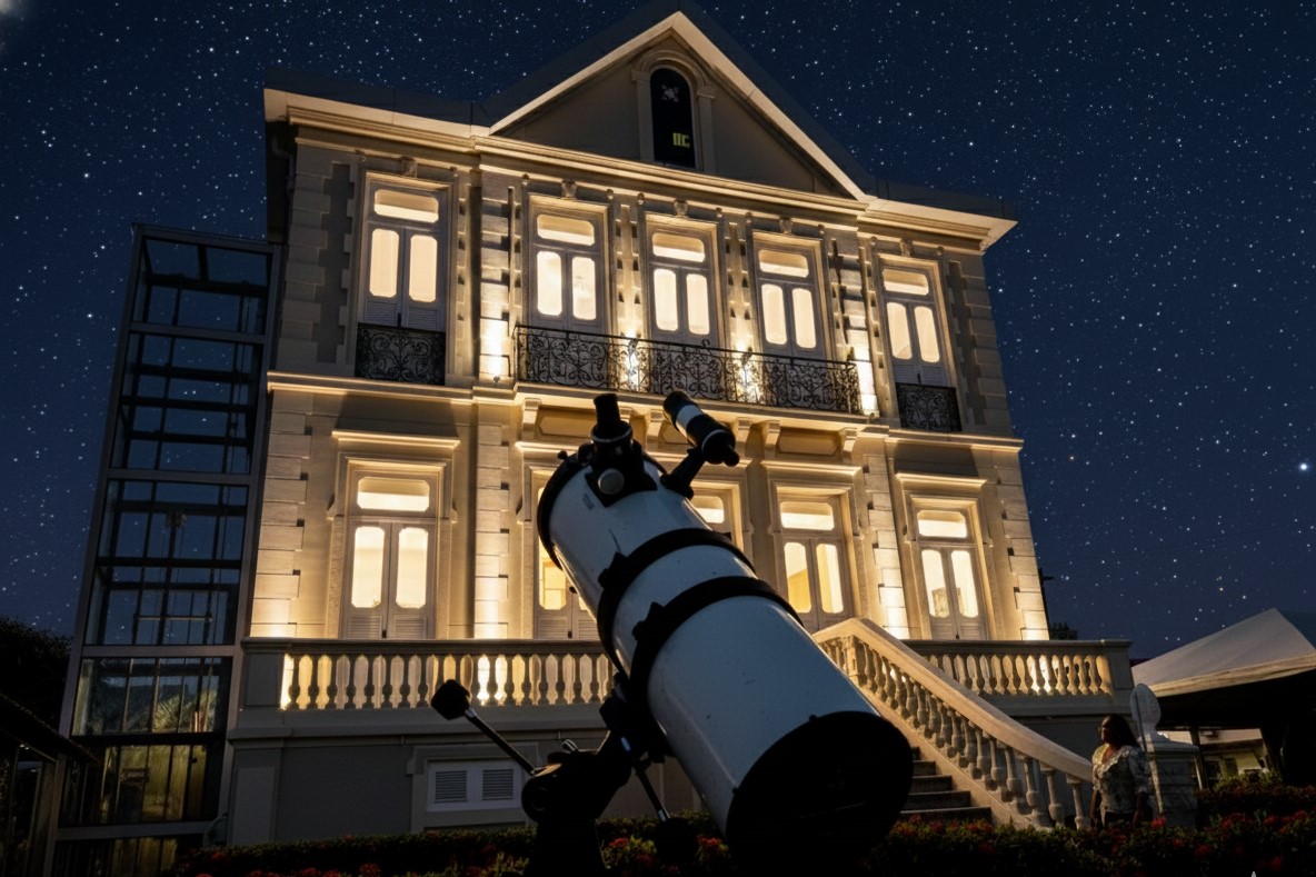 Em primeiro plano na imagem, há um telescópio astronômico apontado para o céu, durante uma atividade de observação astronômica. Ao fundo, vê-se o prédio histórico do Instituto Evandro Chagas, iluminado com luzes amarelas, destacando sua arquitetura clássica com janelas altas, sacadas com grades de ferro e escadaria central.