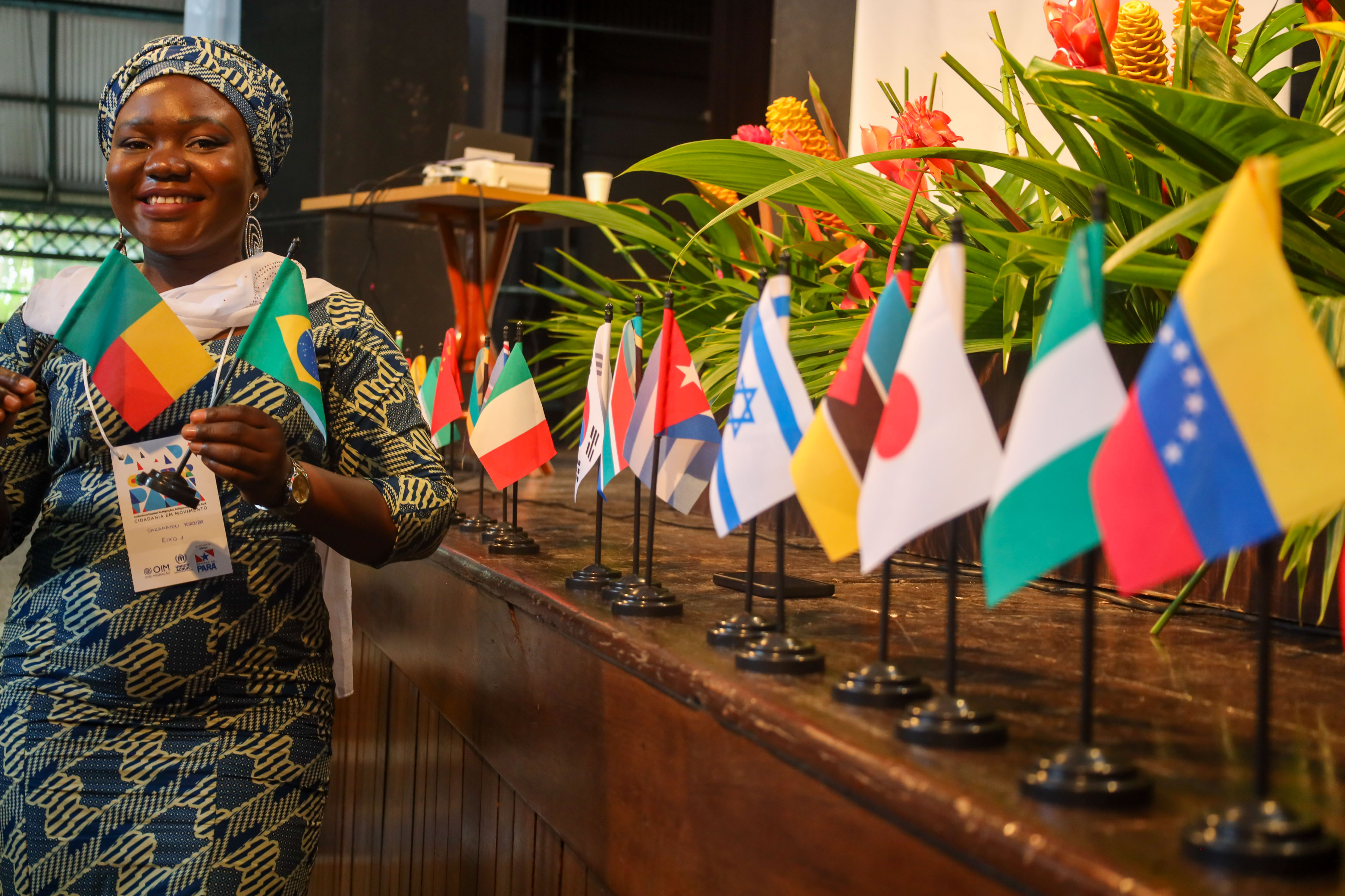Pessoa participante de evento intercultural posa sorrindo segurando as bandeiras do Benin e do Brasil. À sua frente, sobre uma mesa, estão dispostas pequenas bandeiras de diversos países, simbolizando a diversidade cultural e o diálogo entre diferentes nações. Ao fundo, um arranjo de plantas e flores tropicais compõe o cenário do auditório onde ocorreu a programação. A imagem destaca a valorização das mobilidades humanas, da interculturalidade e do acolhimento de migrantes e refugiados.
