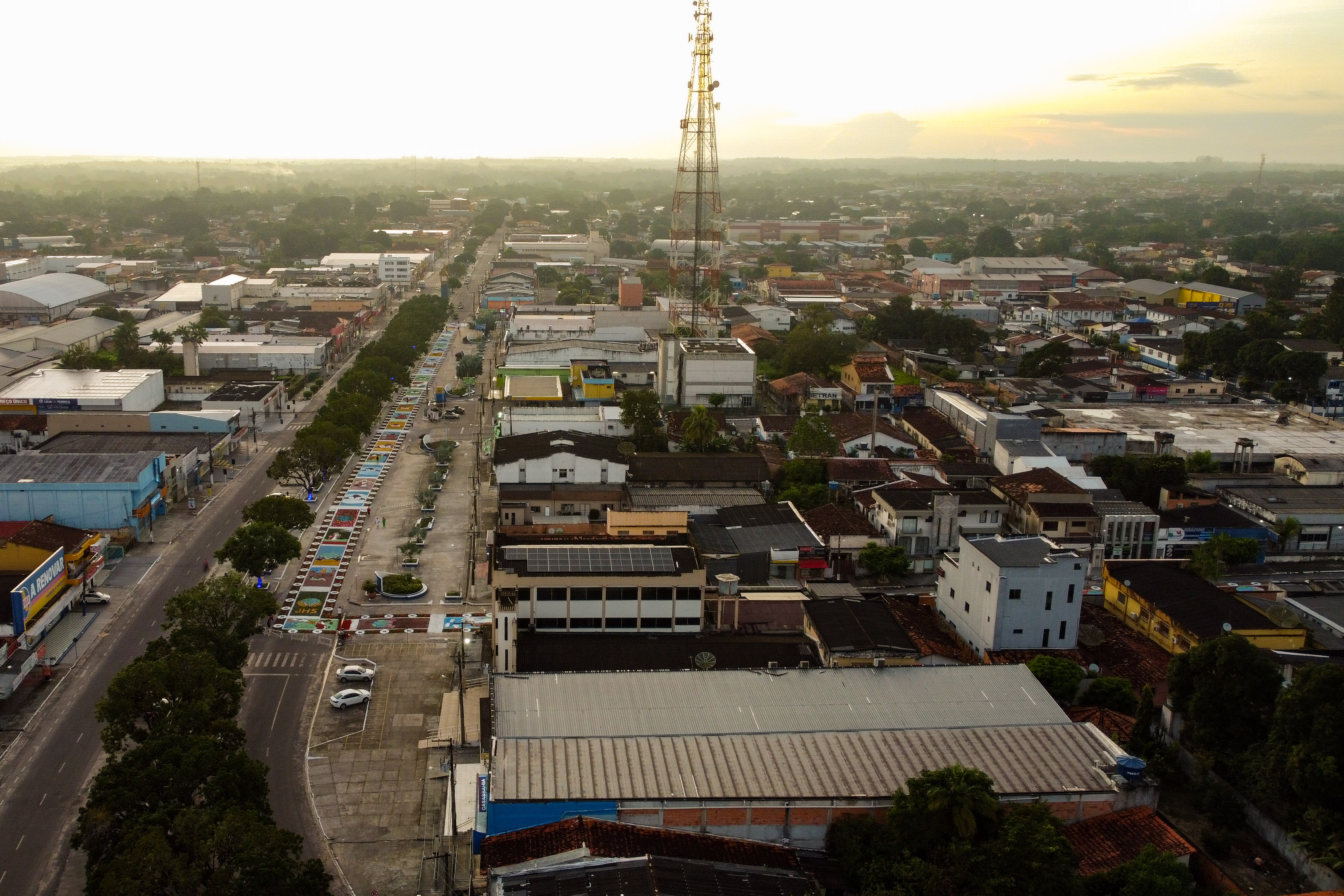 A imagem mostra uma vista área da cidade de Capanema, em um horário de fim de tarde, com um por do sol entre nuvens ao fundo da imagem