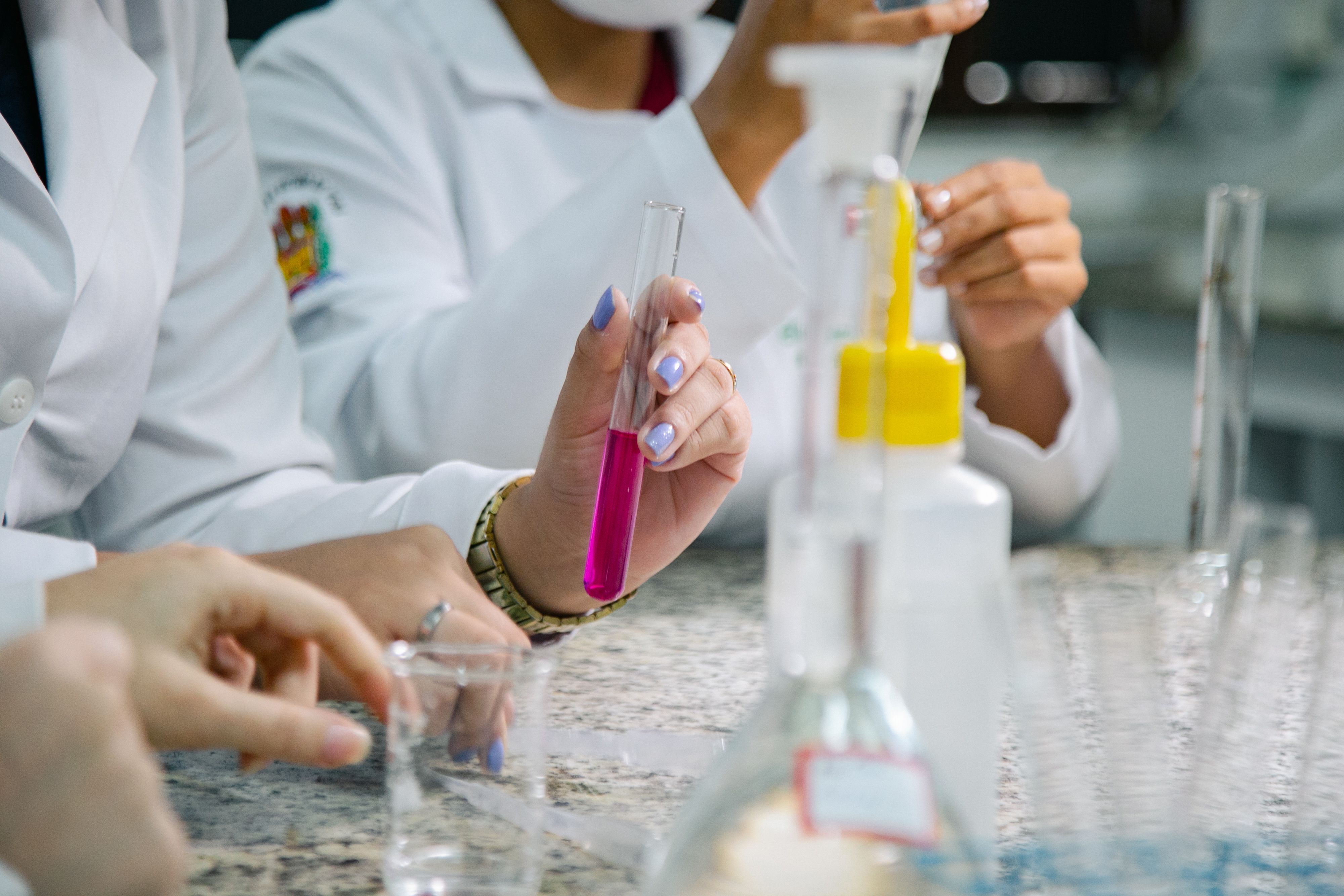 Dois estudantes em um laboratório de química, vestindo jalecos e máscaras, manuseiam vidrarias enquanto uma das mãos segura um tubo de ensaio com líquido rosa sobre uma bancada de granito.