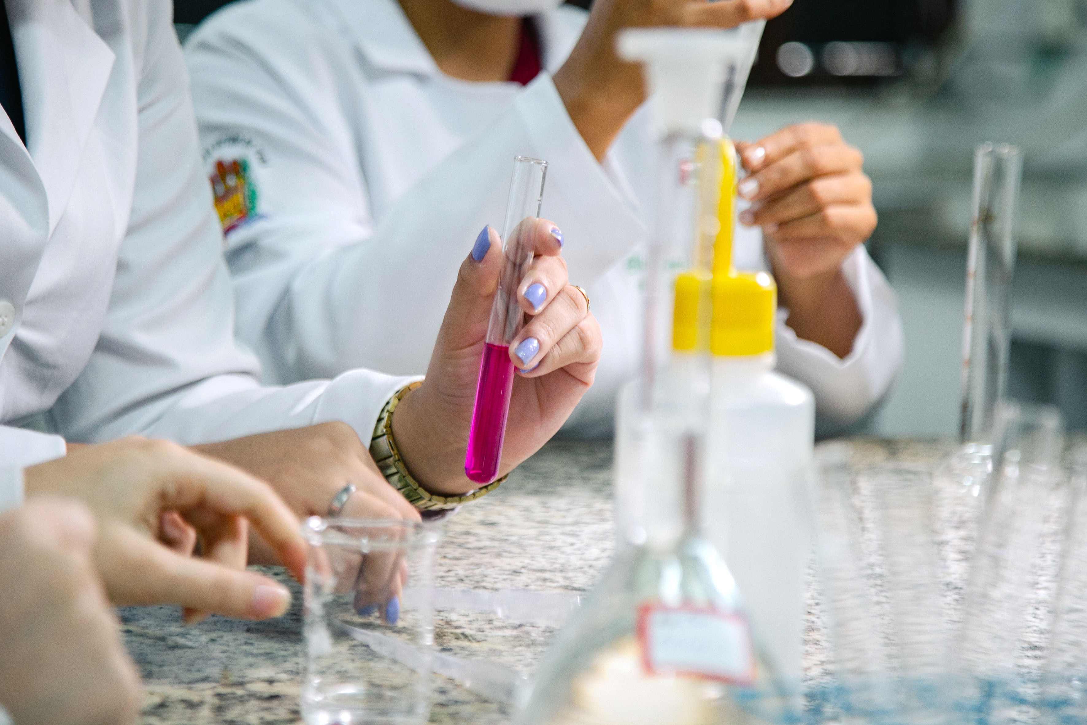Estudantes do curso de Química da Uepa participam de atividade prática em laboratório, manipulando vidrarias e soluções químicas durante experimento. Em primeiro plano, uma estudante segura um tubo de ensaio com líquido de coloração rosa, enquanto outros participantes realizam procedimentos laboratoriais ao fundo. A imagem destaca o ensino prático e a formação científica desenvolvidos na universidade.
