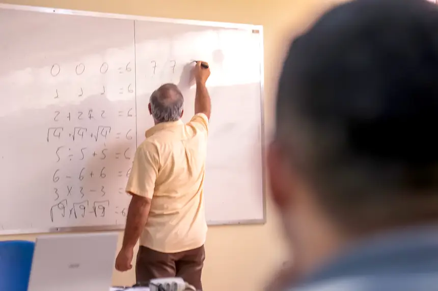 A imagem mostra um professor que ministra aula, escrevendo conteúdos matemáticos no quadro branco, enquanto estudantes acompanham atentamente a explicação em sala de aula.