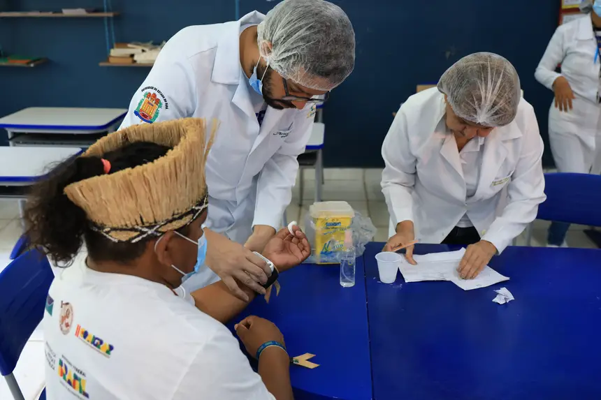A imagem mostra estudantes e profissionais da área da saúde realizam um procedimento de coleta ou avaliação, com o uso de materiais descartáveis e equipamentos de proteção, em uma atividade de caráter educativo e assistencial.